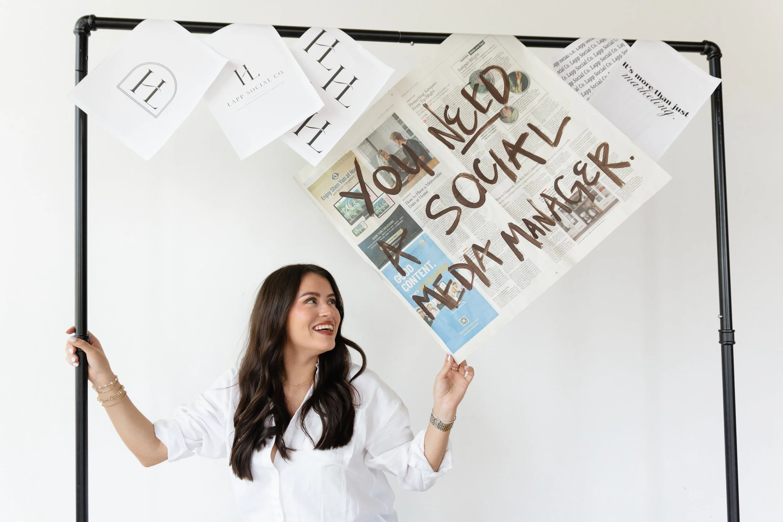 A woman with long brown hair, wearing a white blouse, holding a black pole frame with both hands. The frame has various papers attached, including a newspaper with a message written in large brown letters that says "NEO YOU LOVE YOURSELF AND MEDITATE." The woman is smiling and looking up at the frame.