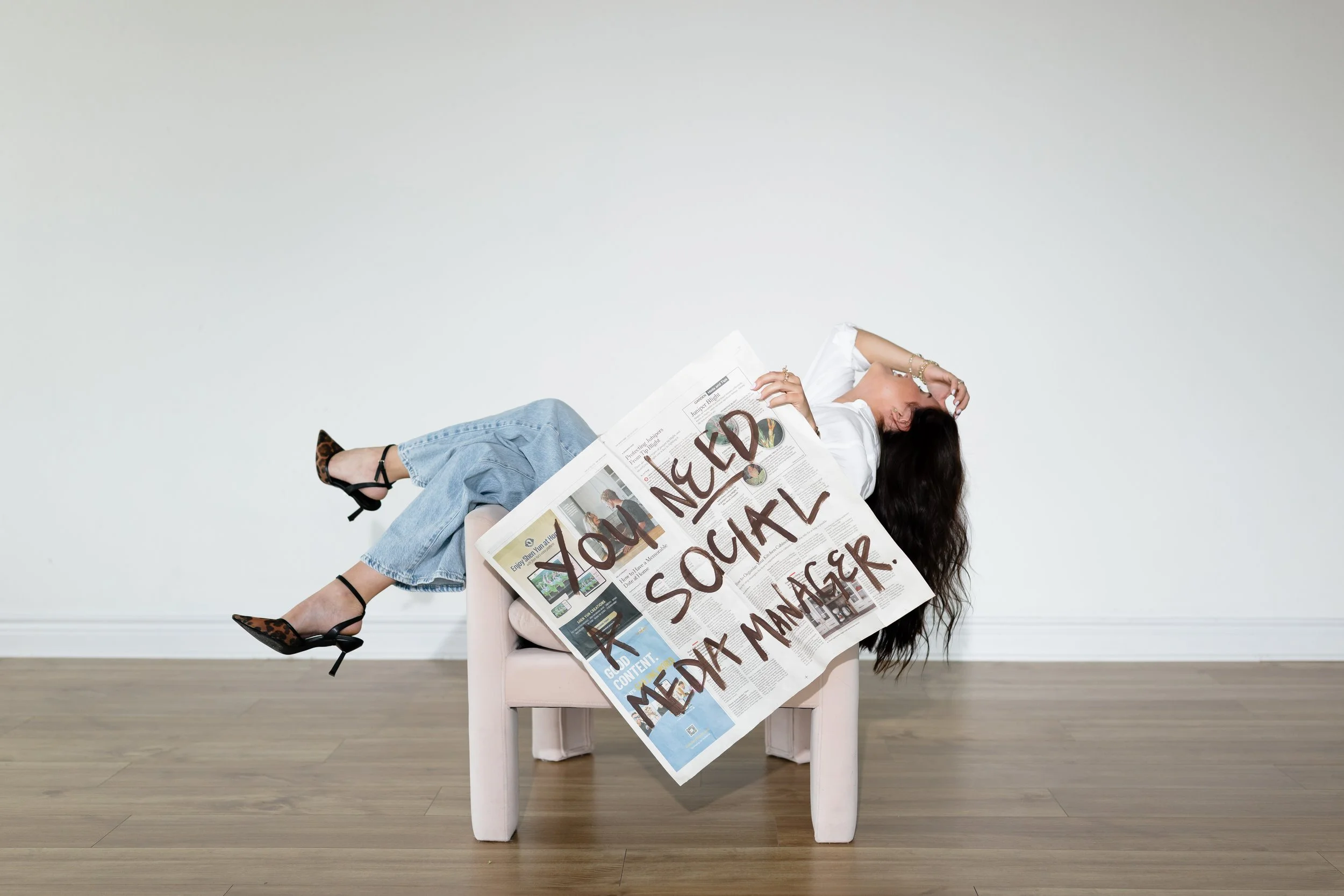 Person lying on a pink chair, reading a large newspaper with headlines written in marker, wearing a white shirt, blue jeans, and leopard print high heels, with long dark hair, against a plain white wall.