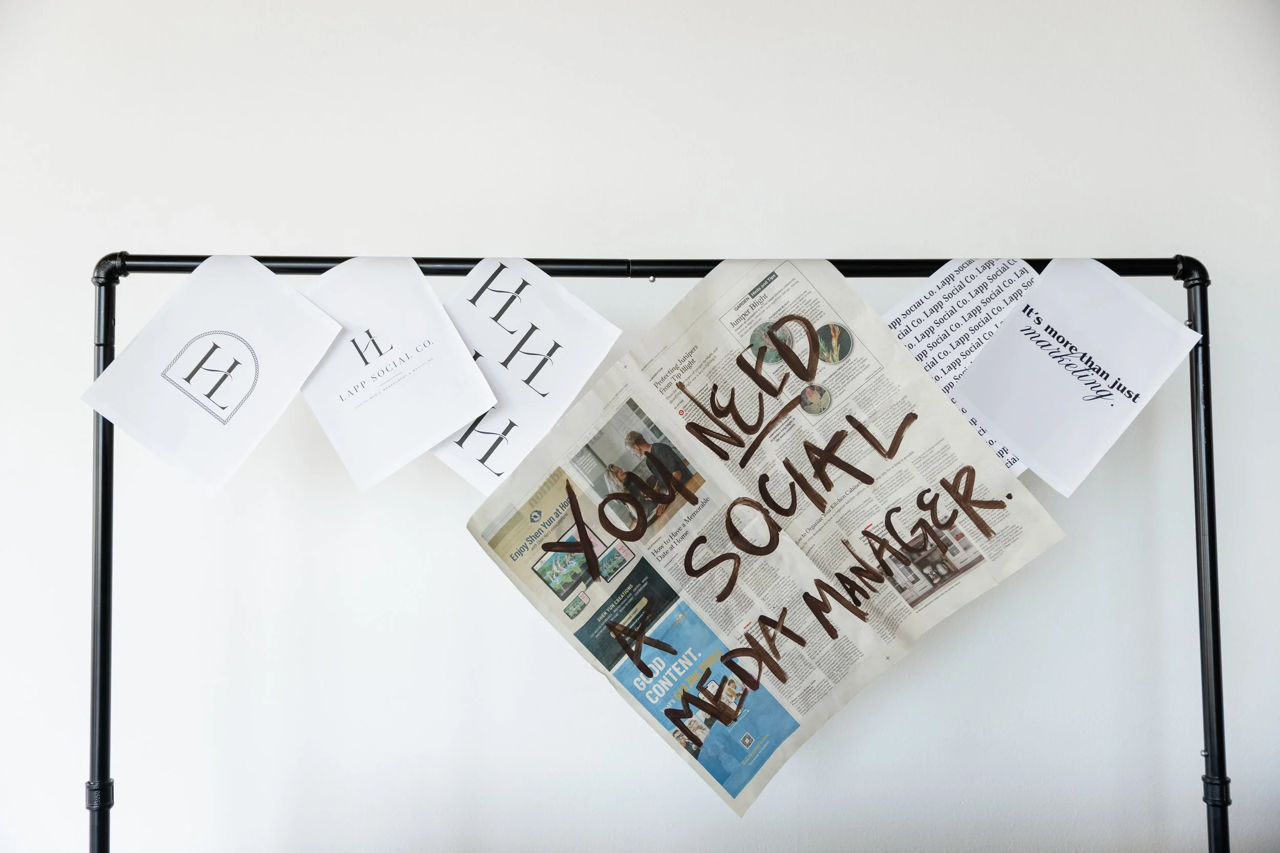 A black metal clothing rack holding several sheets of paper and a folded newspaper, with a handwritten sign in brown marker that reads 'WED NO SAL'.