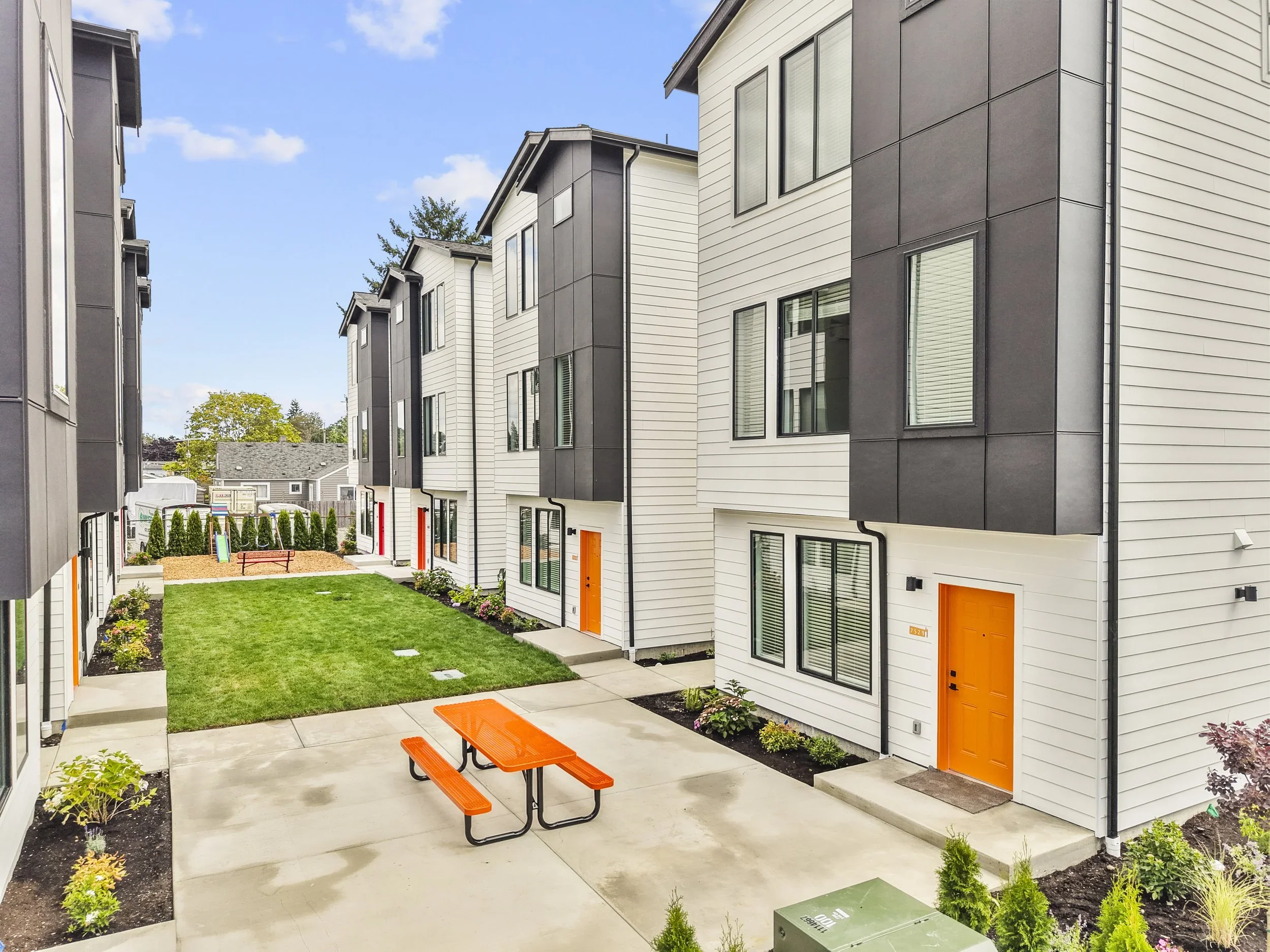 Modern apartment complex with white siding and dark gray panels, featuring bright orange doors. A small courtyard with a green lawn and flower beds is shown, with a concrete patio that includes an orange picnic table. There is a children’s playground with a slide in the background, and a blue sky with some clouds overhead.