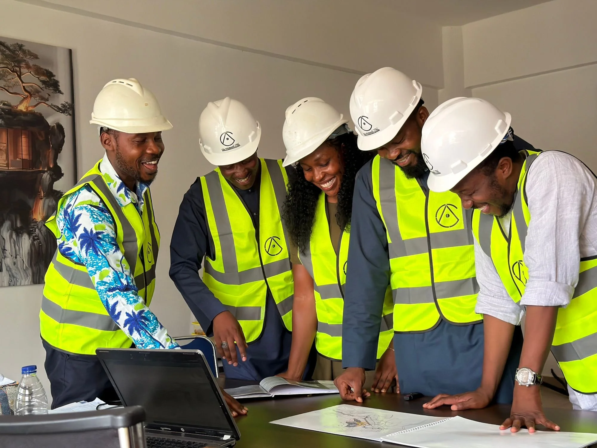 Five people wearing white safety helmets and reflective vests are gathered around a table looking at a large map or document. They appear to be discussing or planning a project.
