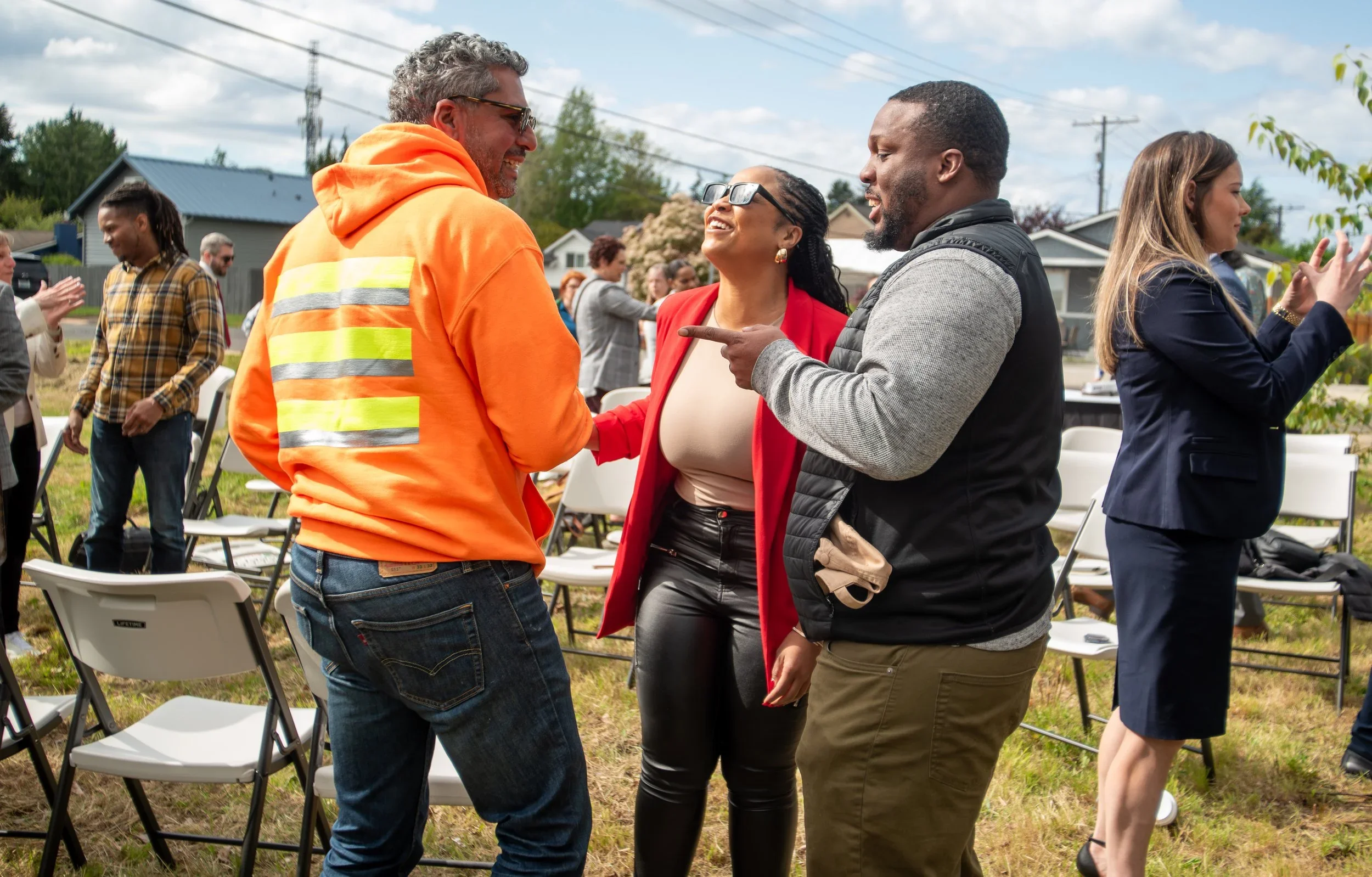 A group of diverse people at an outdoor event, engaging in conversation. The focus is on three individuals in the foreground: a man in an orange safety jacket, a woman in a red blazer, and a man in a gray and black jacket. They are smiling and appear to be having a friendly discussion. Other attendees are visible in the background, seated and standing, with chairs and houses also in view under a partly cloudy sky.