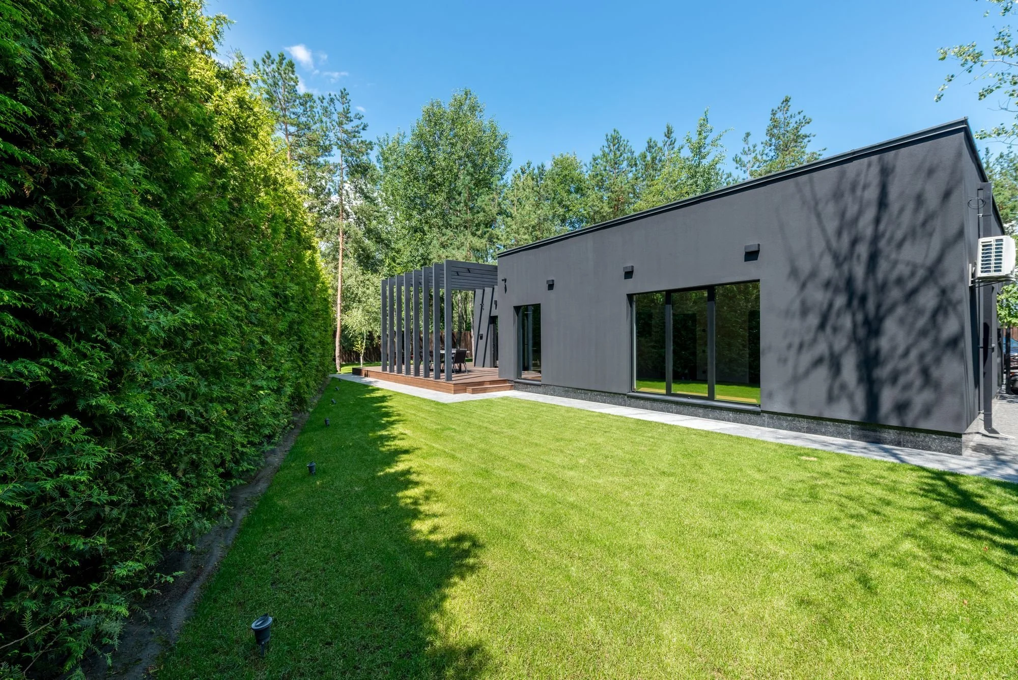 Modern grey house with large glass windows and a small wooden deck, surrounded by a well-manicured lawn and tall green trees on a sunny day.