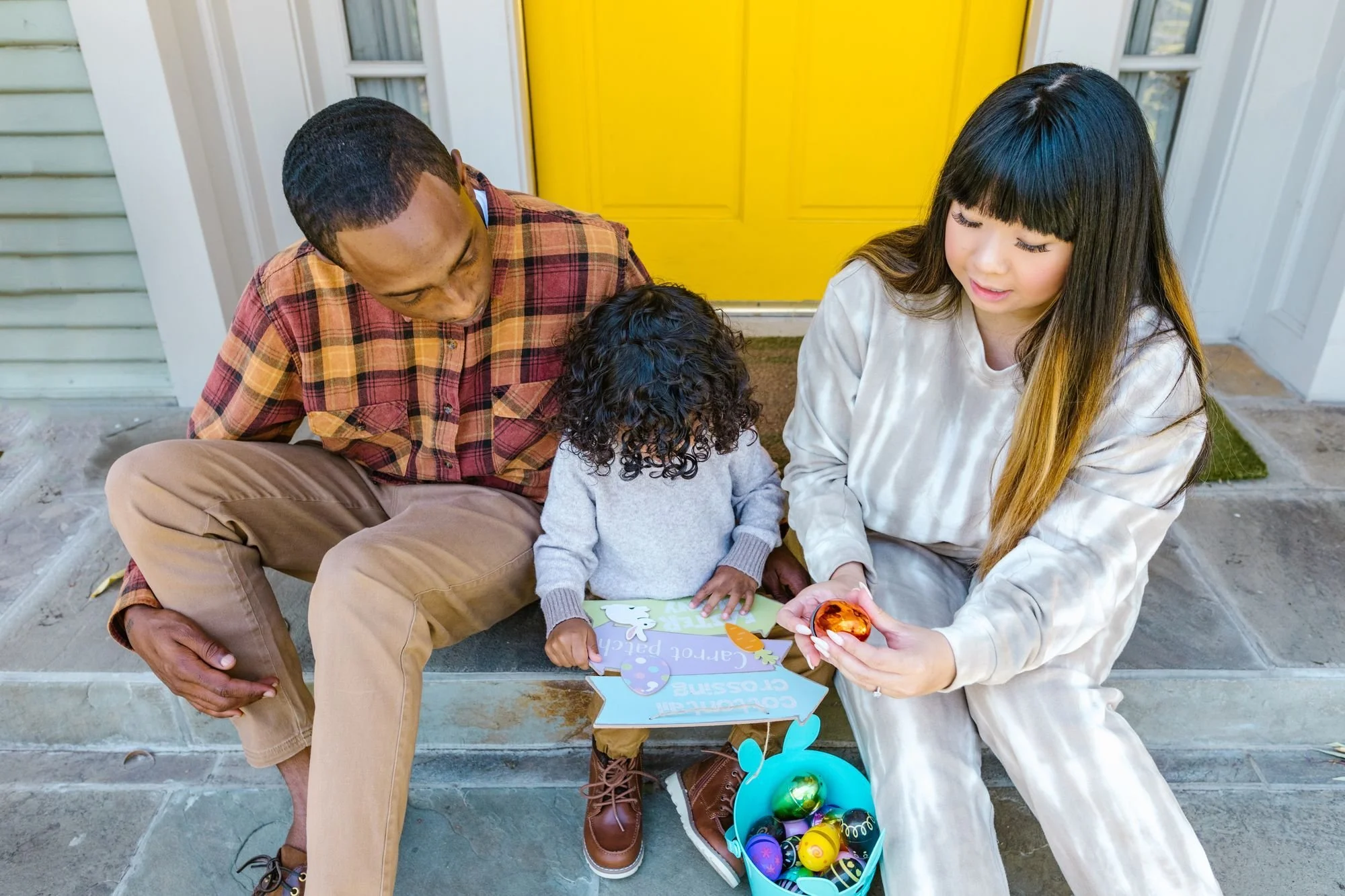 Family of three on front porch celebrating Easter. Woman hands child decorated eggs, man looks on. Child with curly hair writes on greeting card, basket of colorful Easter eggs nearby.