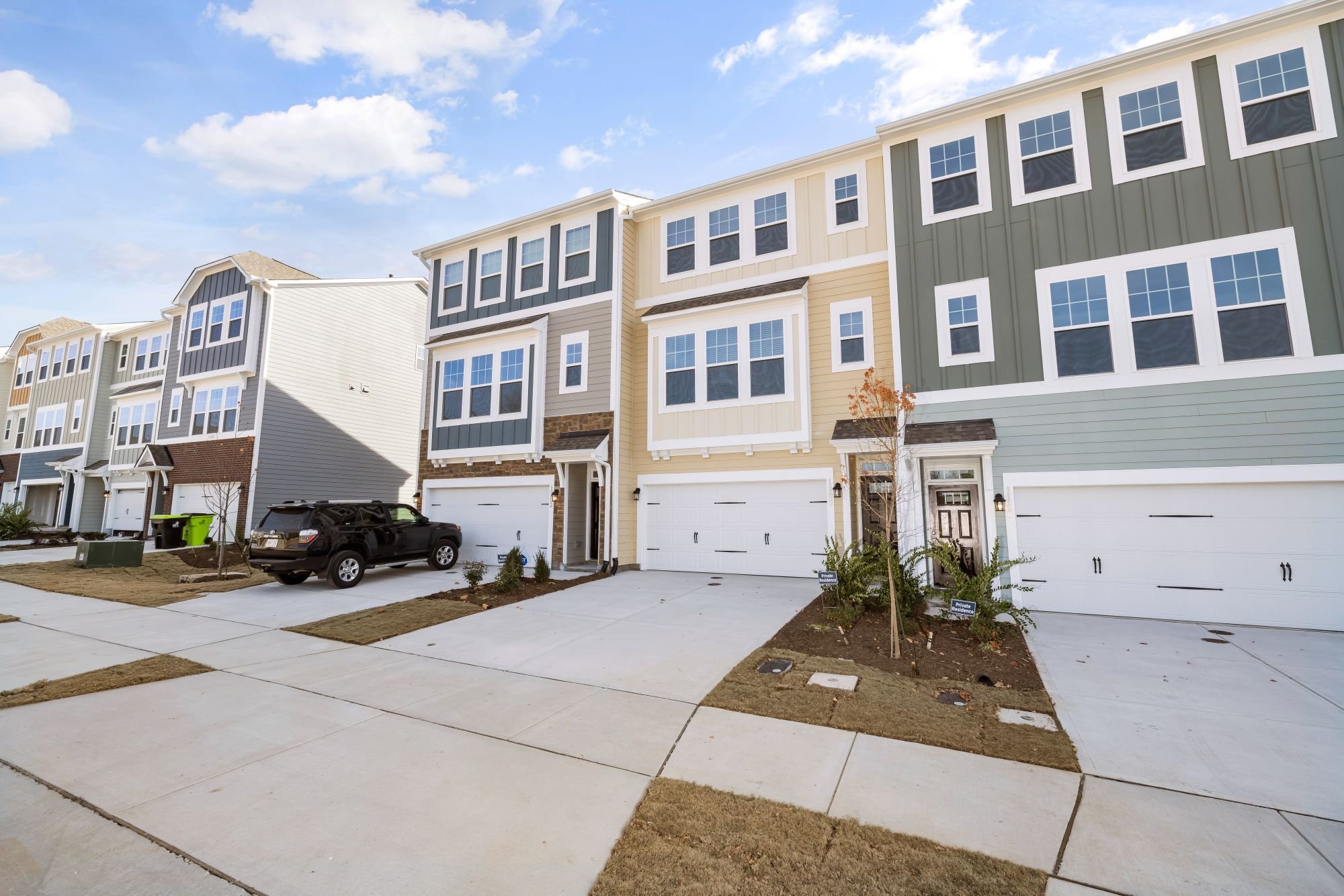 Row of modern townhouses with multiple floors, large windows, and attached garages. Cars are parked in front of the houses, and small trees and landscaping are present. The sky is partly cloudy.