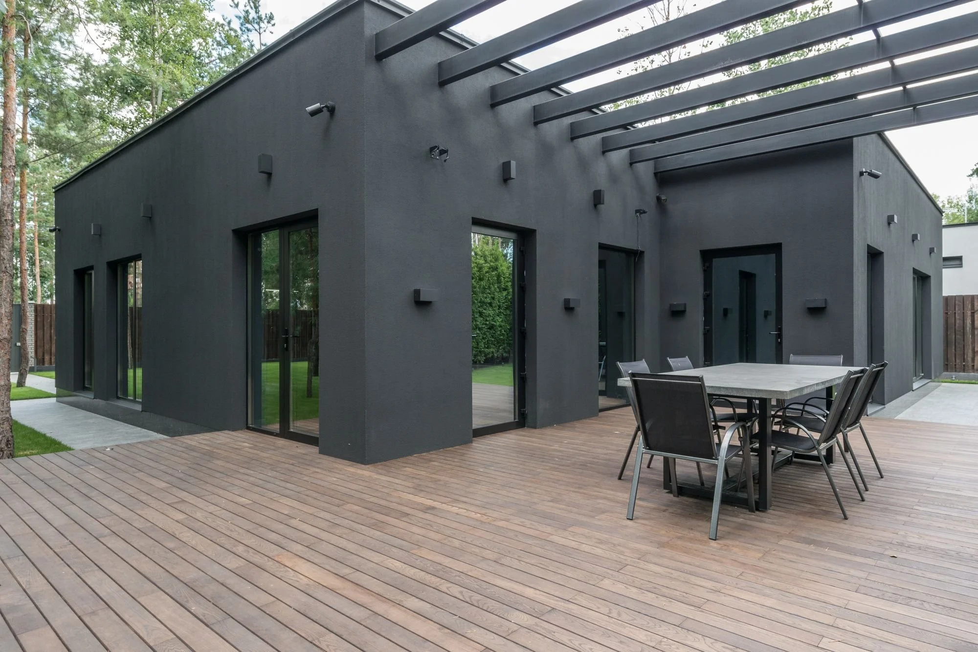 Modern black house with large sliding glass doors, an outdoor dining table with chairs, a wooden deck, and surrounded by tall trees.