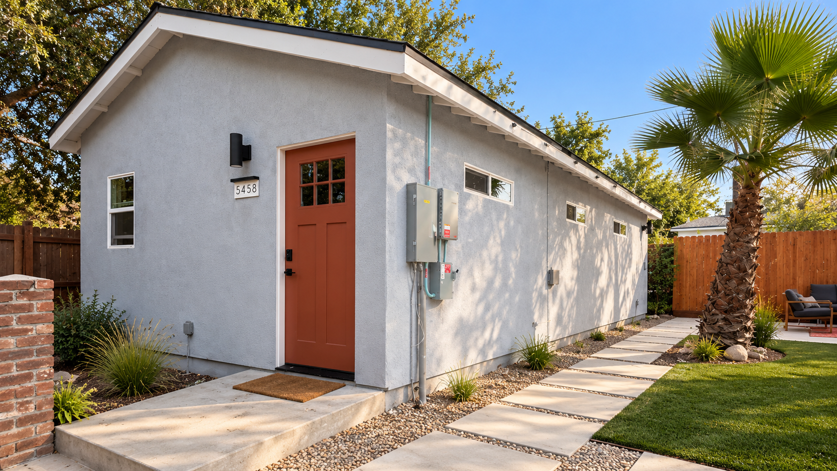 Modern gray house with a red door, small windows, and landscaping including a palm tree, gravel pathway, and outdoor seating area, sunny day with blue sky.