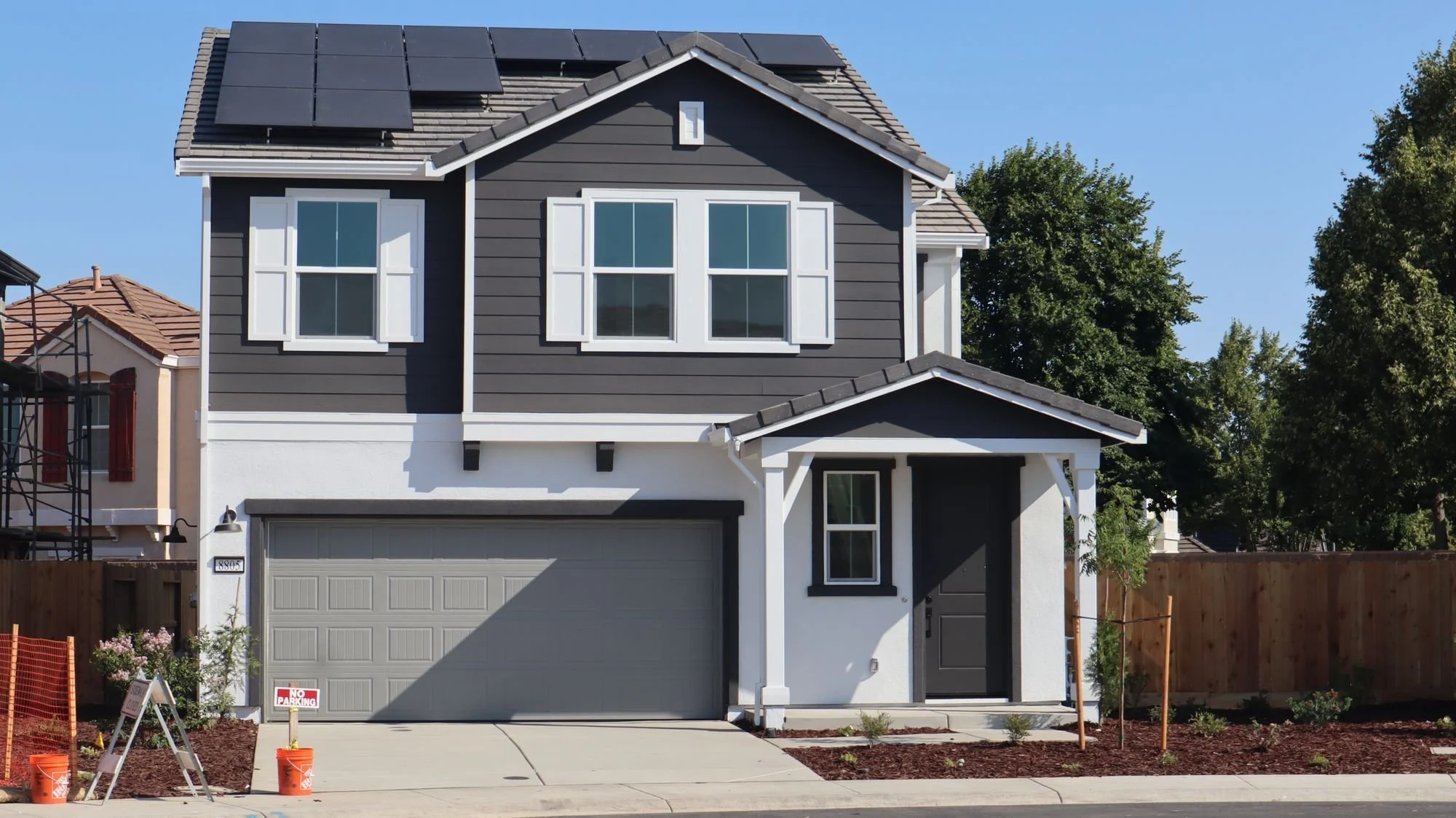 A modern two-story house with dark gray siding, white trim, and a light gray garage door. Solar panels are installed on the roof. There is a small front yard with a freshly planted garden and construction materials nearby.