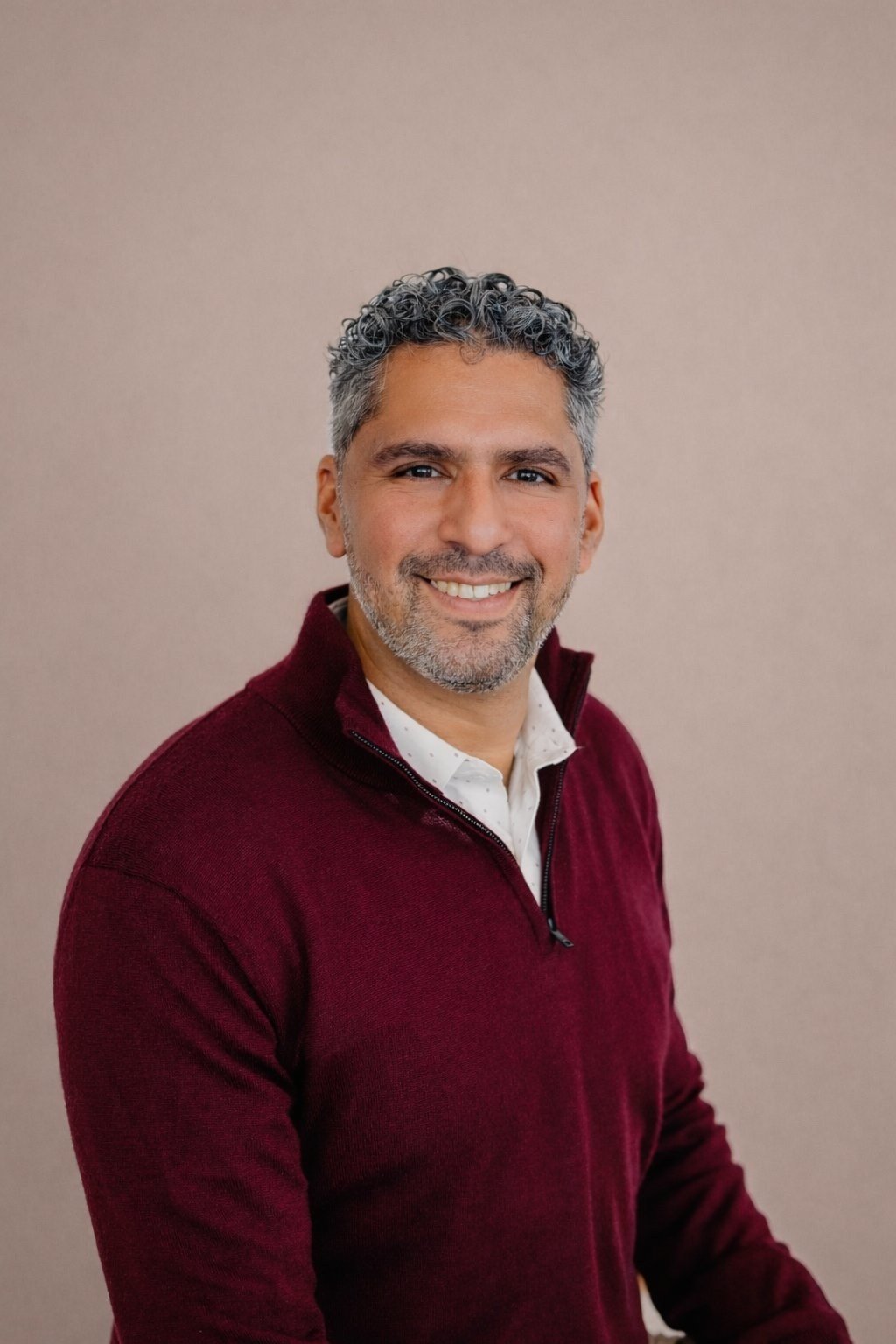 A smiling man with curly dark hair and a beard, wearing a burgundy sweater and a white collared shirt, standing against a plain beige background.