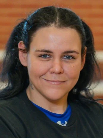 A young woman with dark hair styled in pigtails, smiling with a brick wall in the background.