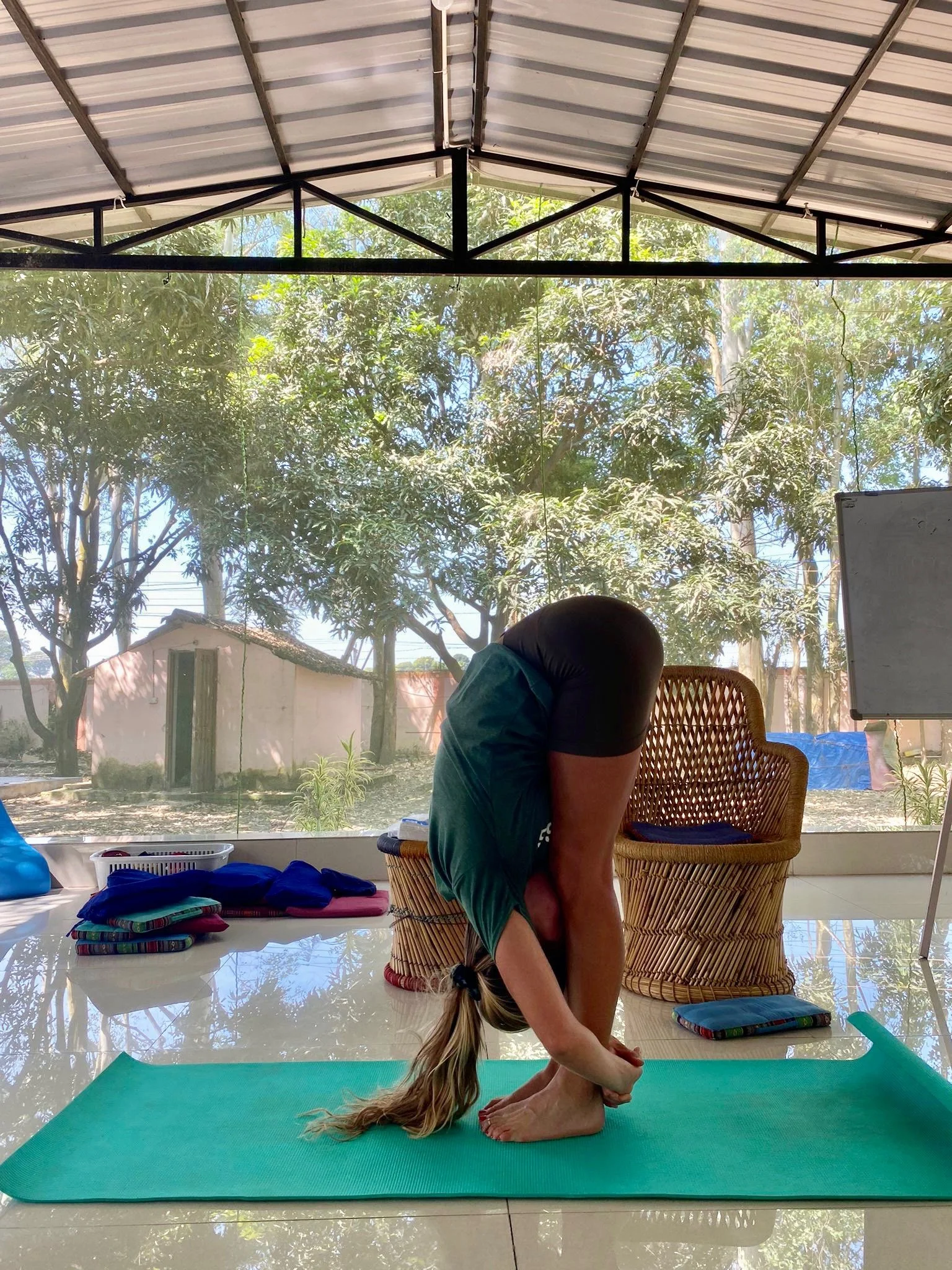 A woman with long blonde hair practicing yoga indoors near large windows with a view of trees and a small building outside. She is in a forward bend pose on a teal yoga mat, wearing a dark t-shirt and shorts. Various folded clothes or towels are on the floor beside her.