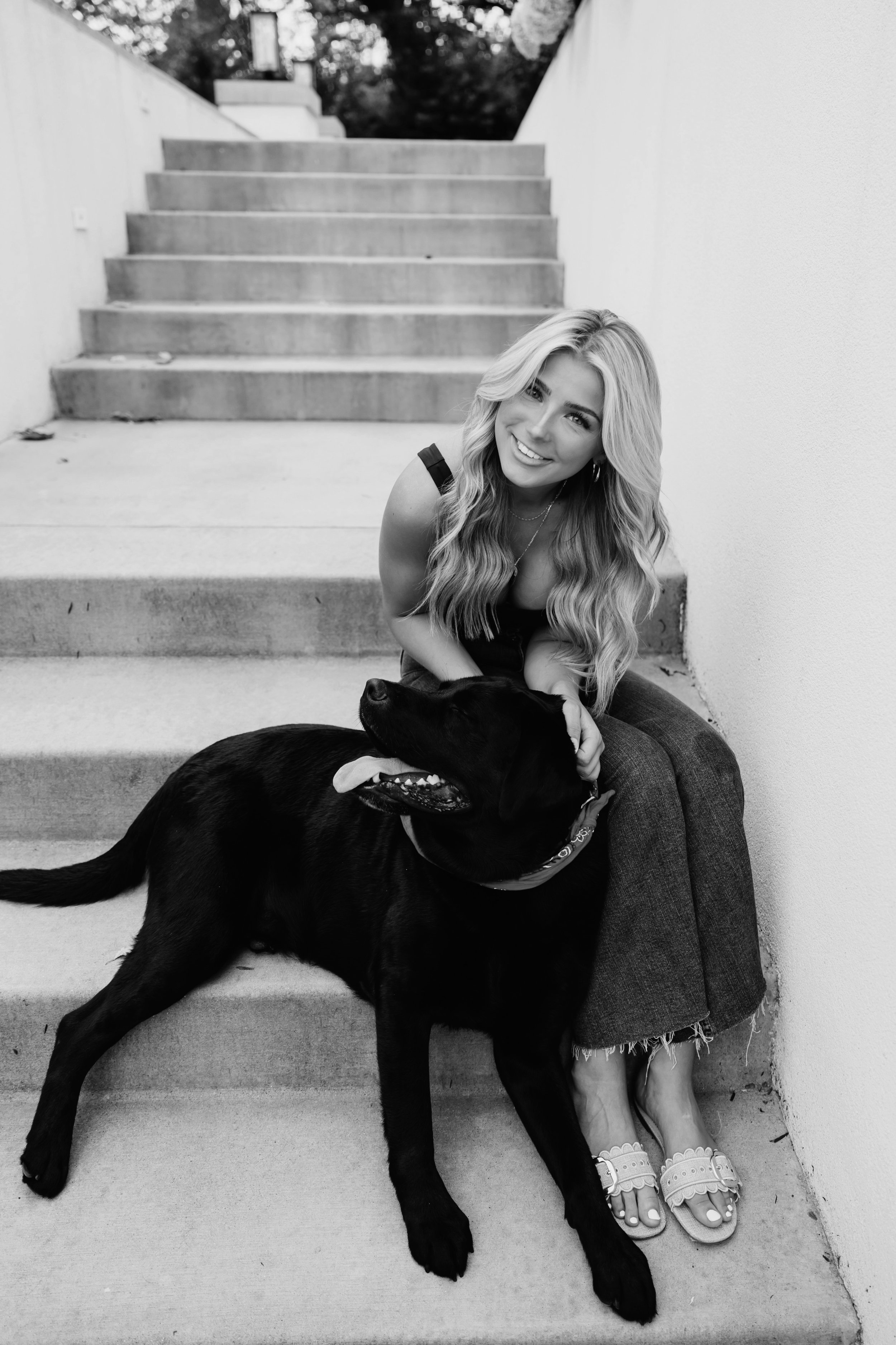 A woman with long wavy hair sitting on outdoor stairs, smiling at the camera, holding a large black dog, both appearing happy and relaxed.