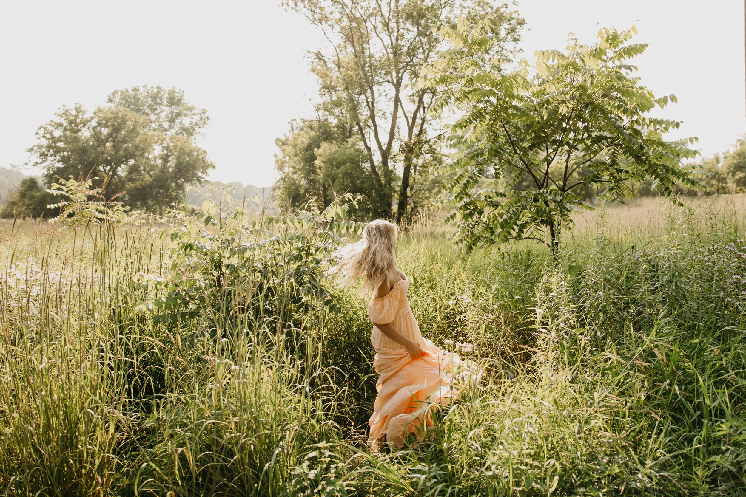 A young girl with long blonde hair wearing a flowing peach dress sitting in tall grass in a sunlit field with trees in the background.