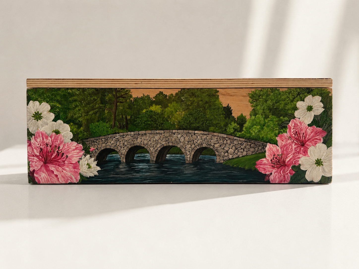 Painted wooden box illustrating a stone bridge over a river with greenery and pink and white flowers on the sides.