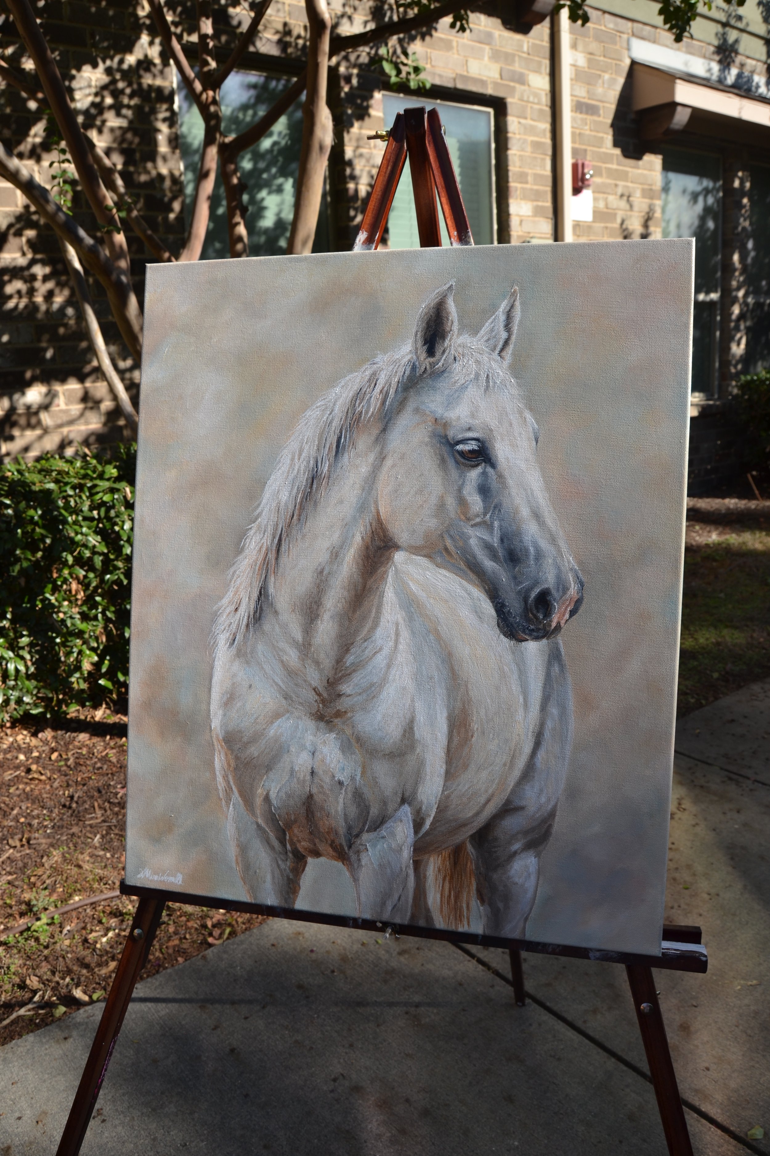 A detailed painting of a white horse with a gray mane, shown on an easel outdoors in front of a brick building and green foliage.