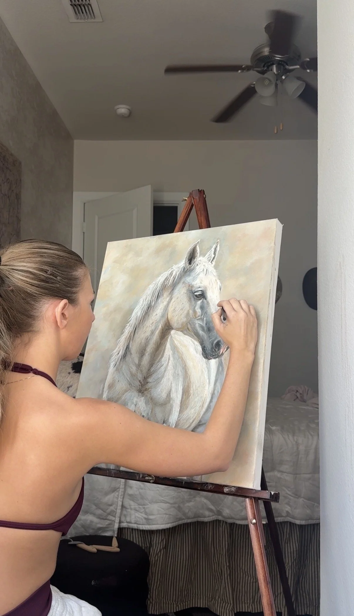 A woman with blonde hair tied back is painting a realistic portrait of a white horse on a canvas in a room with white walls, a ceiling fan, and a door in the background.