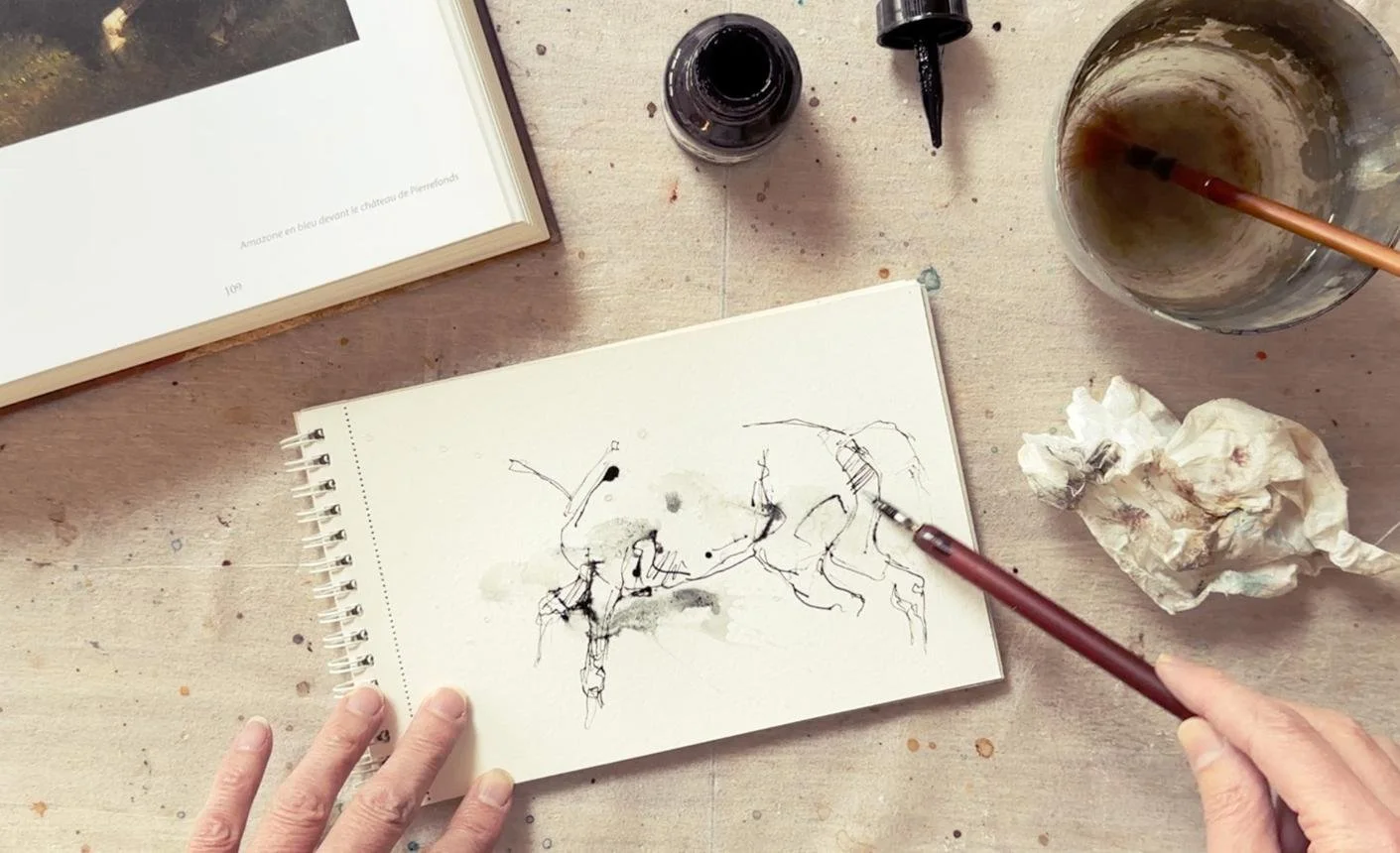 A person's hands working on a sketchbook with a pen. The sketch contains abstract ink drawings. Around the workspace are a cup with brown liquid and a paintbrush, an open ink bottle, crumpled tissue paper, and a book with a photo of a bird on the page.