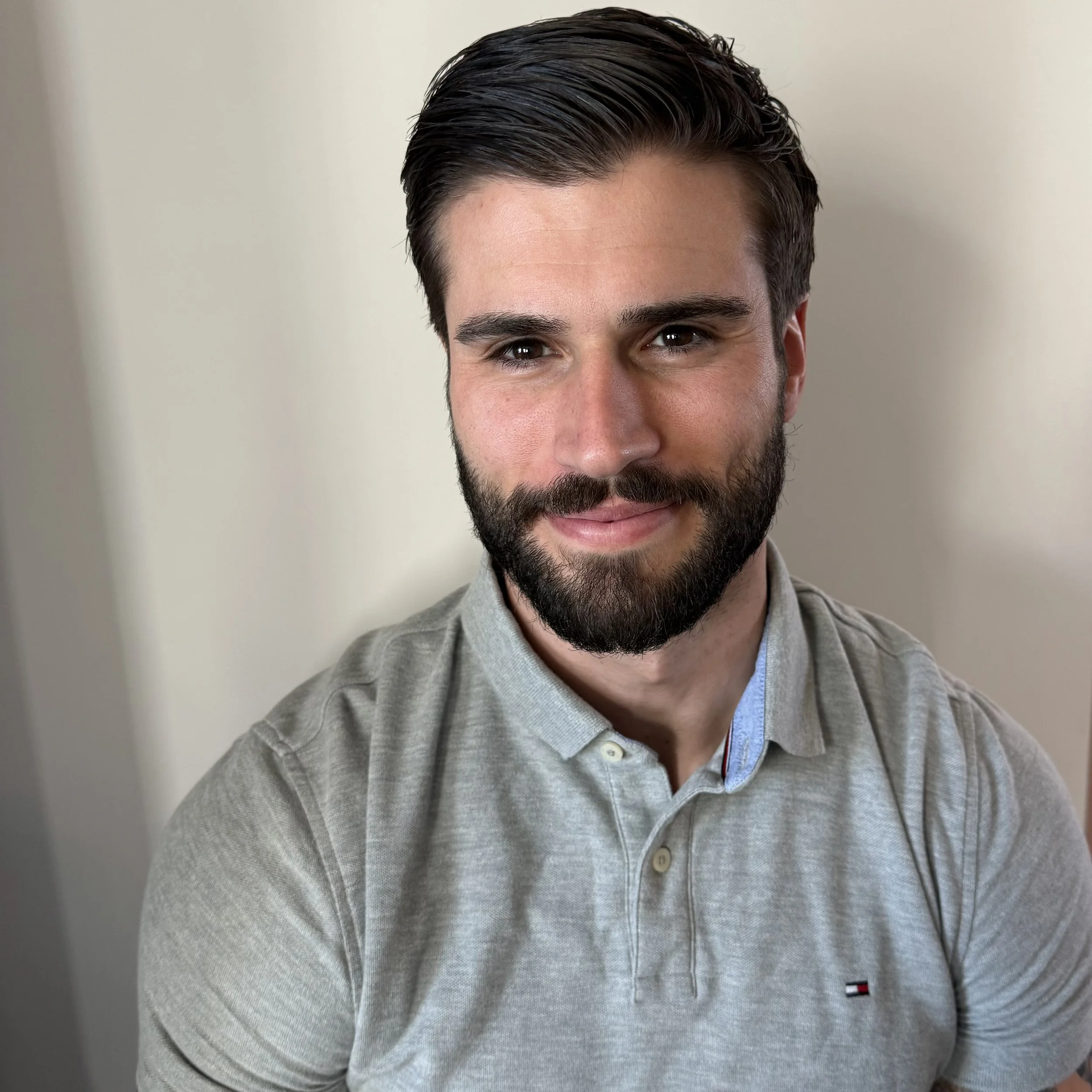 Close-up of a man with dark, styled hair, a trimmed beard, wearing a light grey collared shirt with a small Tommy Hilfiger logo on the chest, against a plain off-white background.
