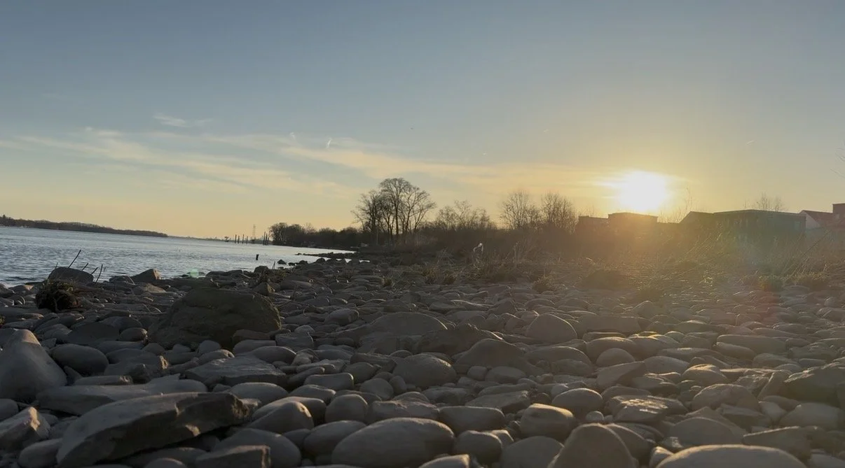 Pebble-covered shoreline with a river, trees, and houses in the distance as the sun sets on a clear evening.