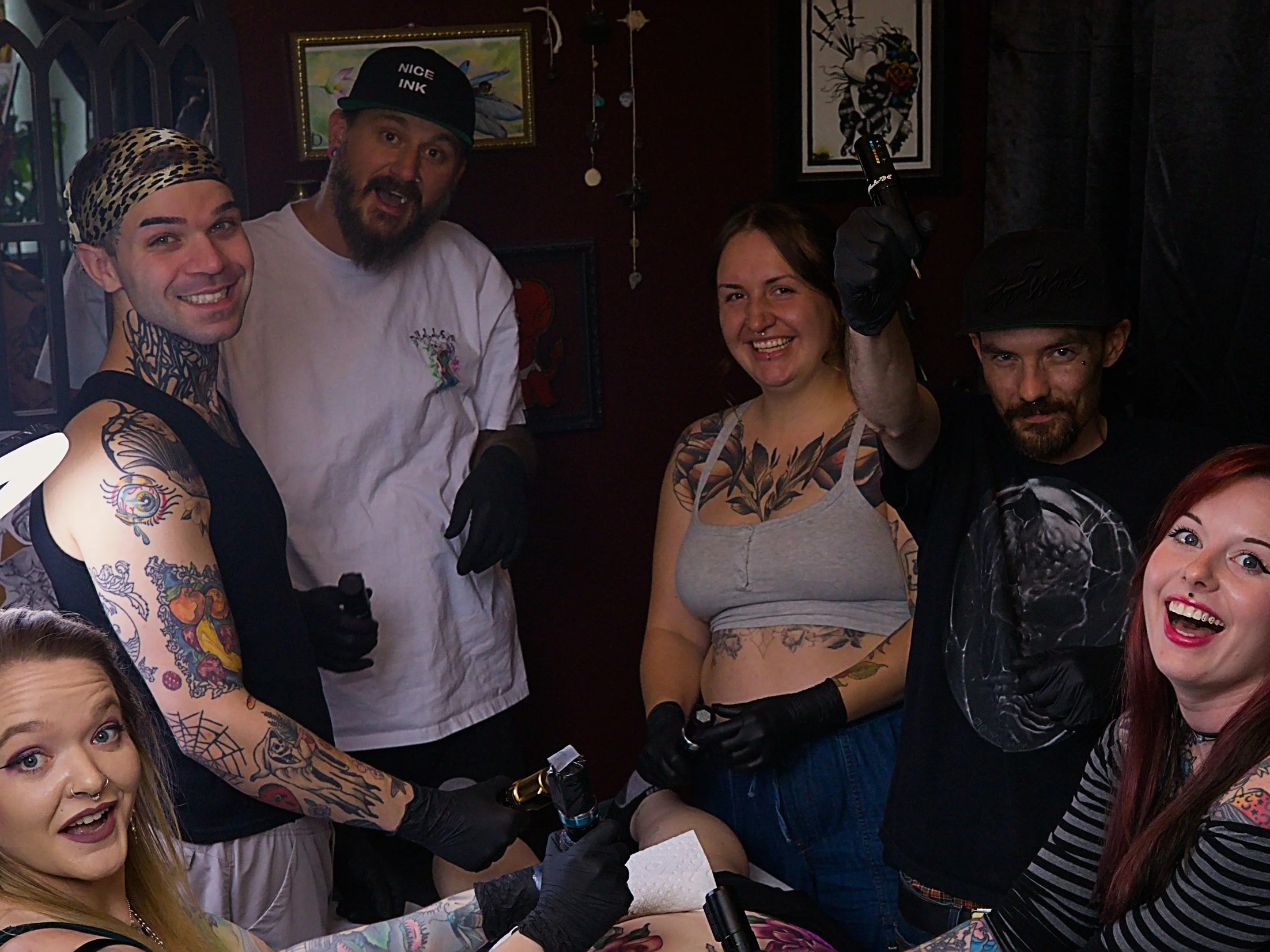Group of tattooed people celebrating at a tattoo studio, with some smiling and others holding tattoo machines.