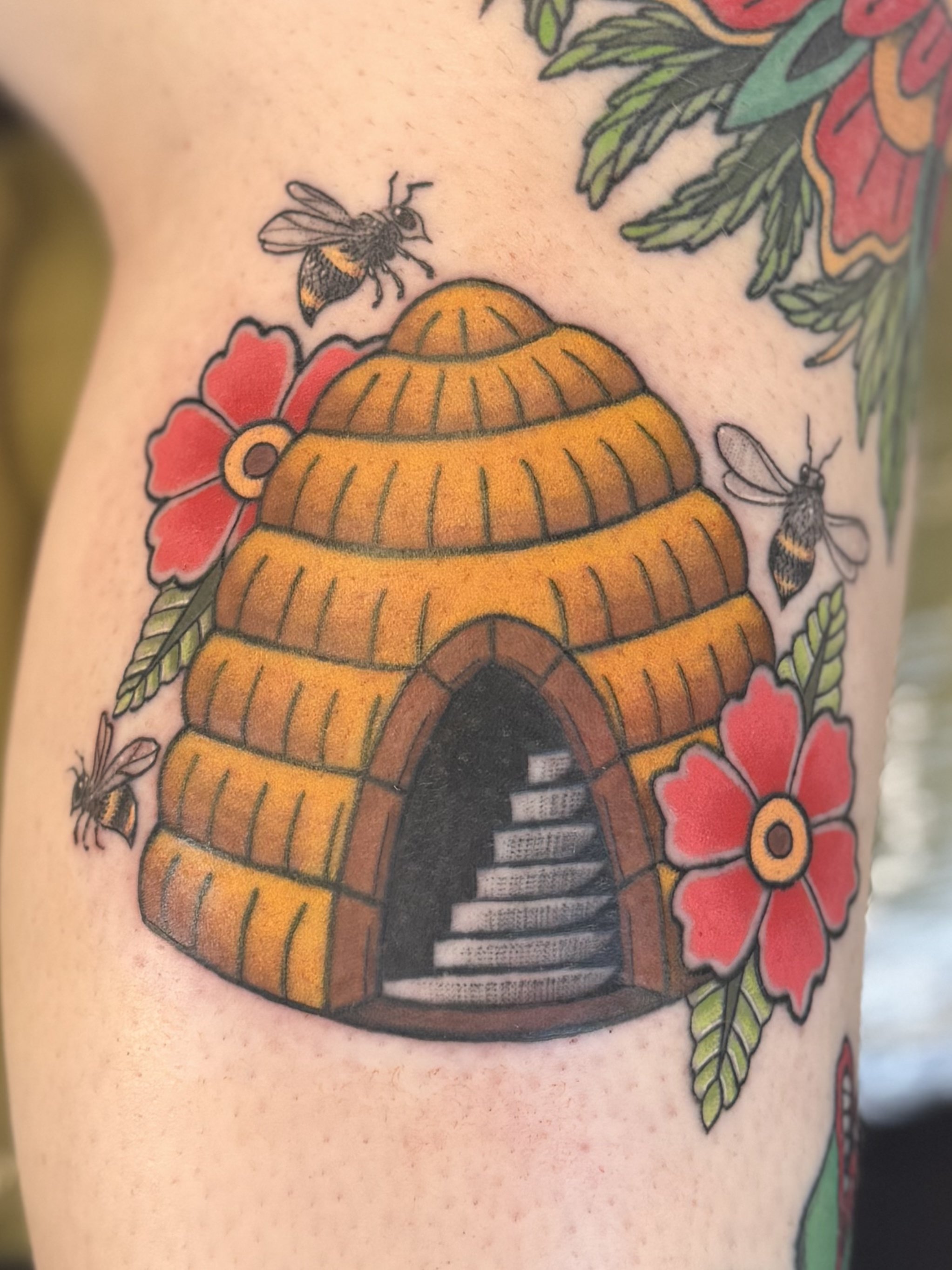 A colorful tattoo of a beehive with two bees flying near it, surrounded by red flowers and green leaves.