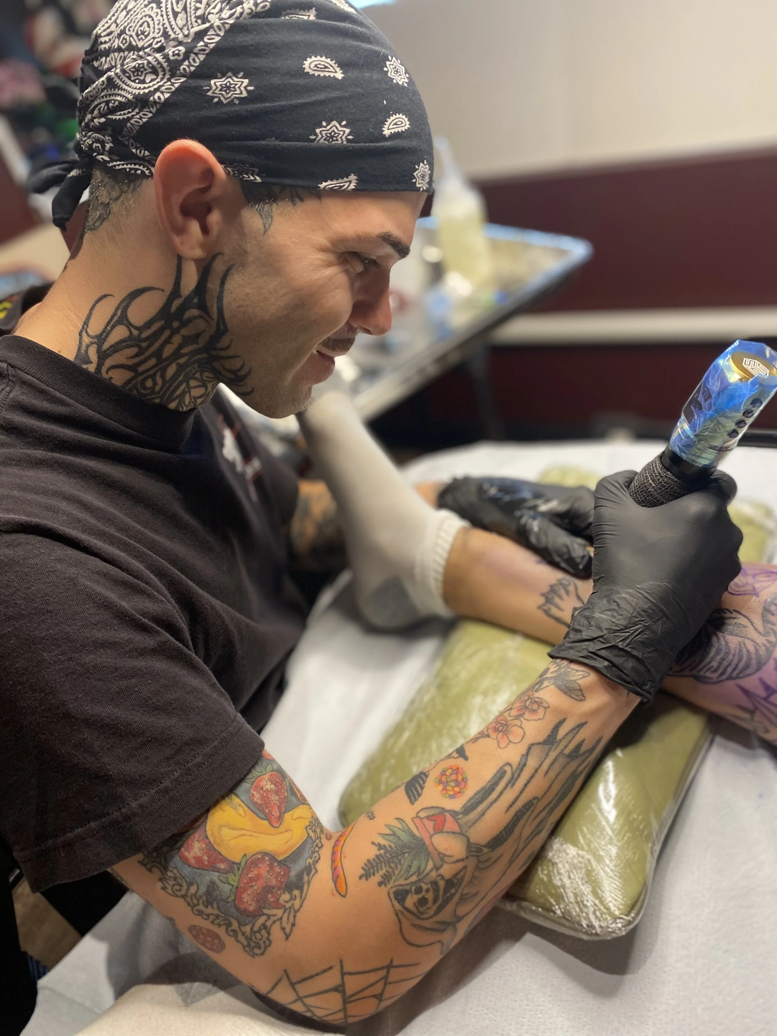 A tattooed man wearing a bandana and black gloves is getting a colorful tattoo on his forearm. The tattoo features flowers, animals, and abstract designs.