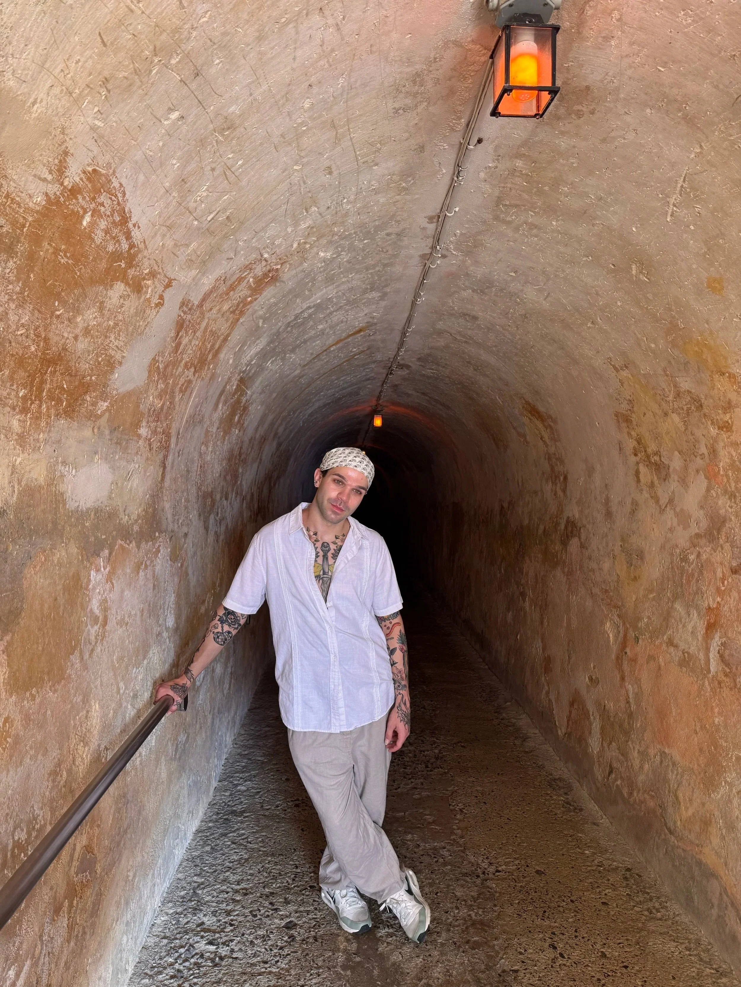 A man with tattoos on his arms and neck, wearing white pants, a white shirt, white sneakers, and a headscarf, standing in a tunnel with textured, weathered orange-brown walls and a black ceiling. There is a handrail on one side, and above him, there are two orange lights and a black fixture on the ceiling.