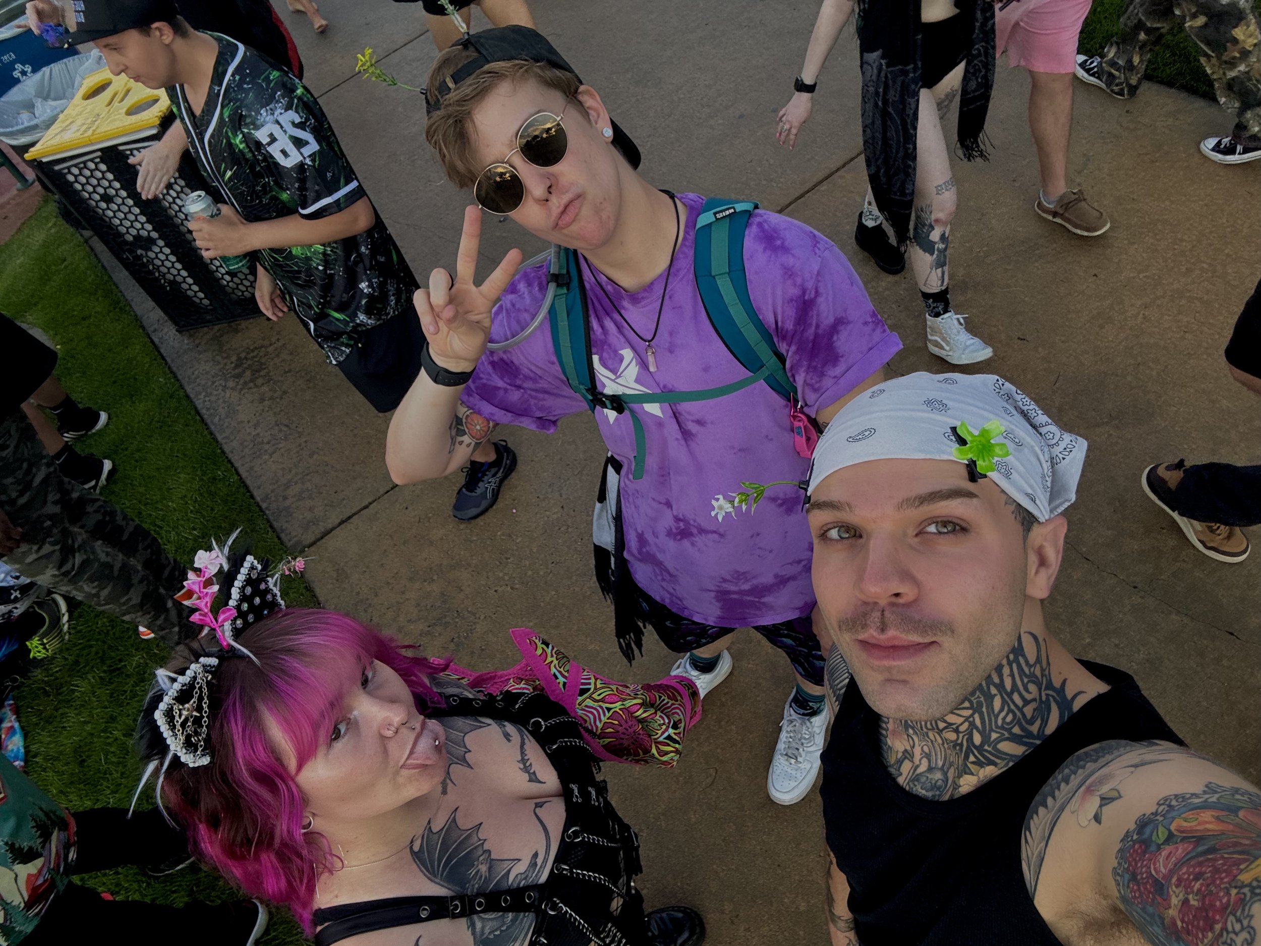 A group of young people at an outdoor event taking a selfie, with two individuals making peace signs. The group includes a person with pink hair, tattoos, and colorful clothing, and others with different hairstyles and accessories.