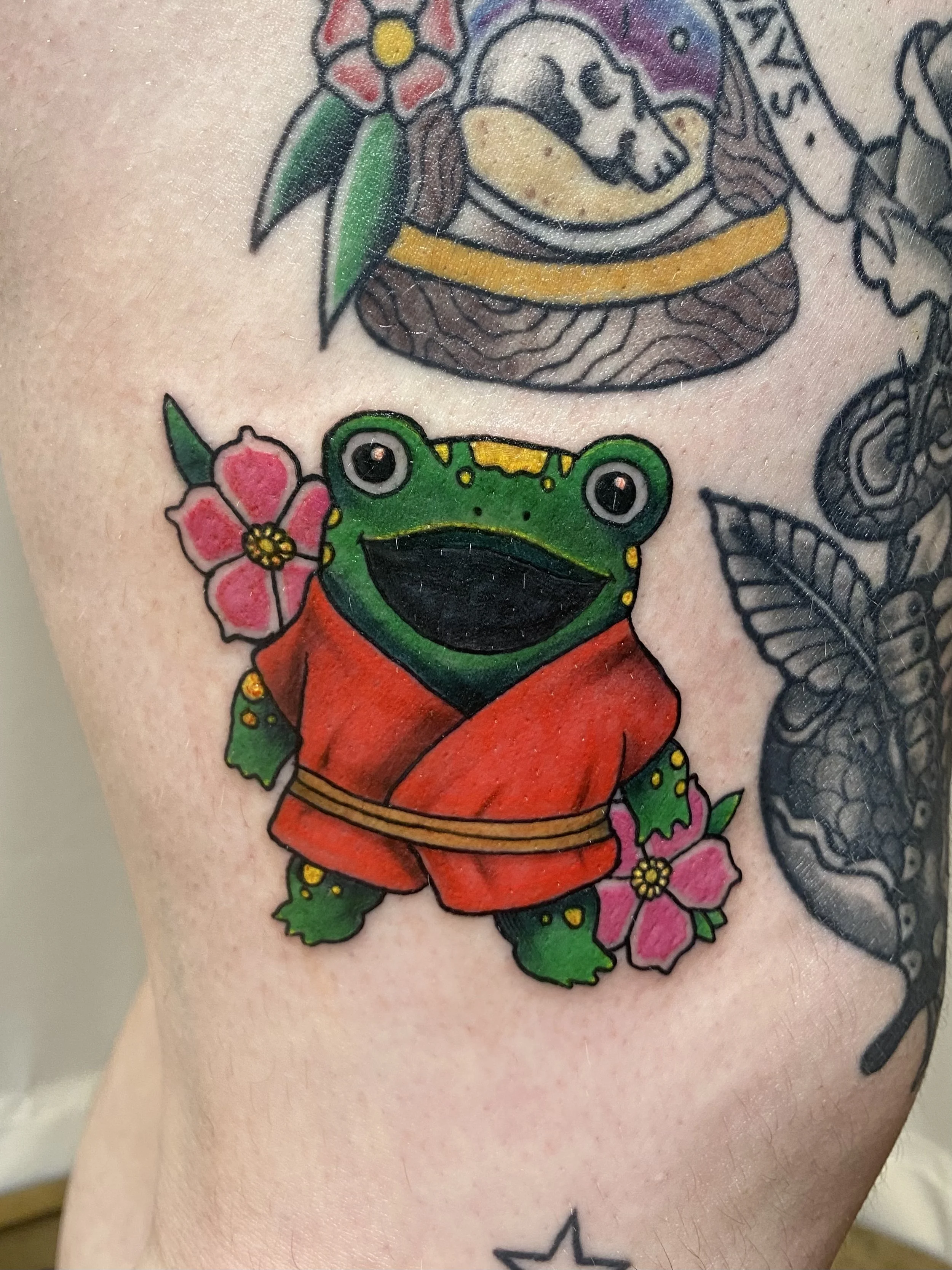 A tattoo of a frog wearing a red kimono, with pink flowers and green leaves around it. The frog has large, round eyes and a big smiling mouth.