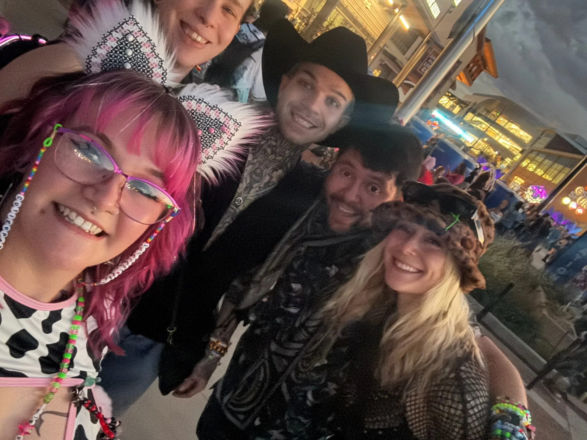 Group of friends smiling at a carnival or amusement park in the evening, some wearing cowboy hats and colorful jewelry.