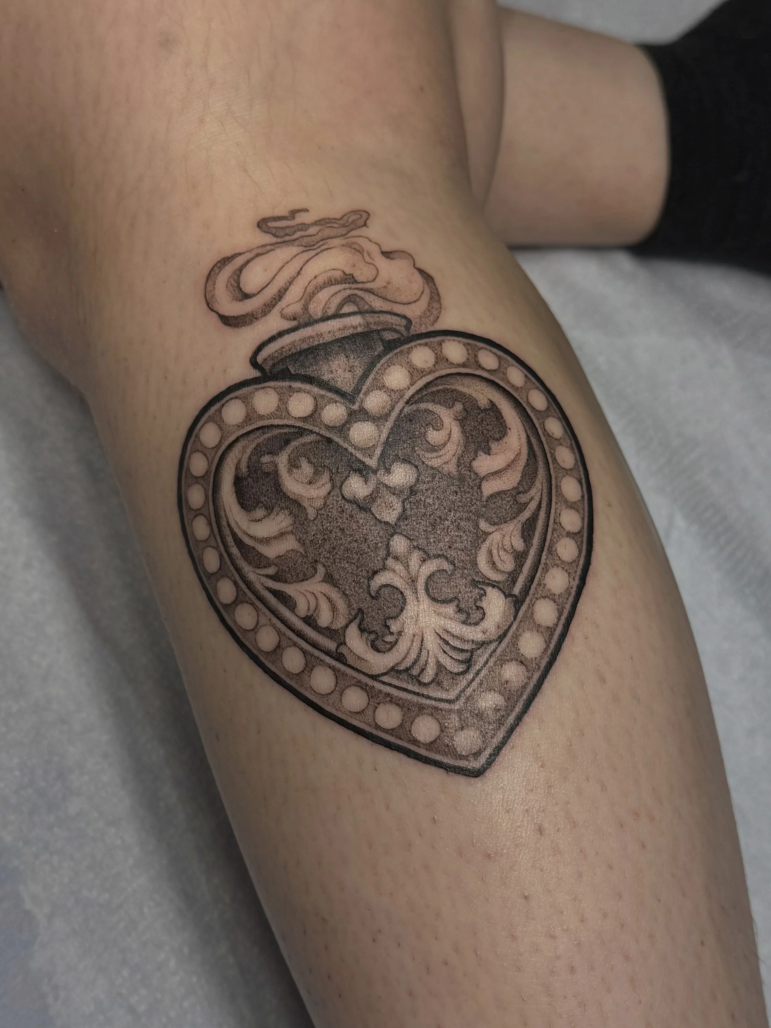 Close-up of a tattoo on a person's upper arm featuring a heart with intricate patterns and an ornamental design inside, with additional decorative elements above the heart.
