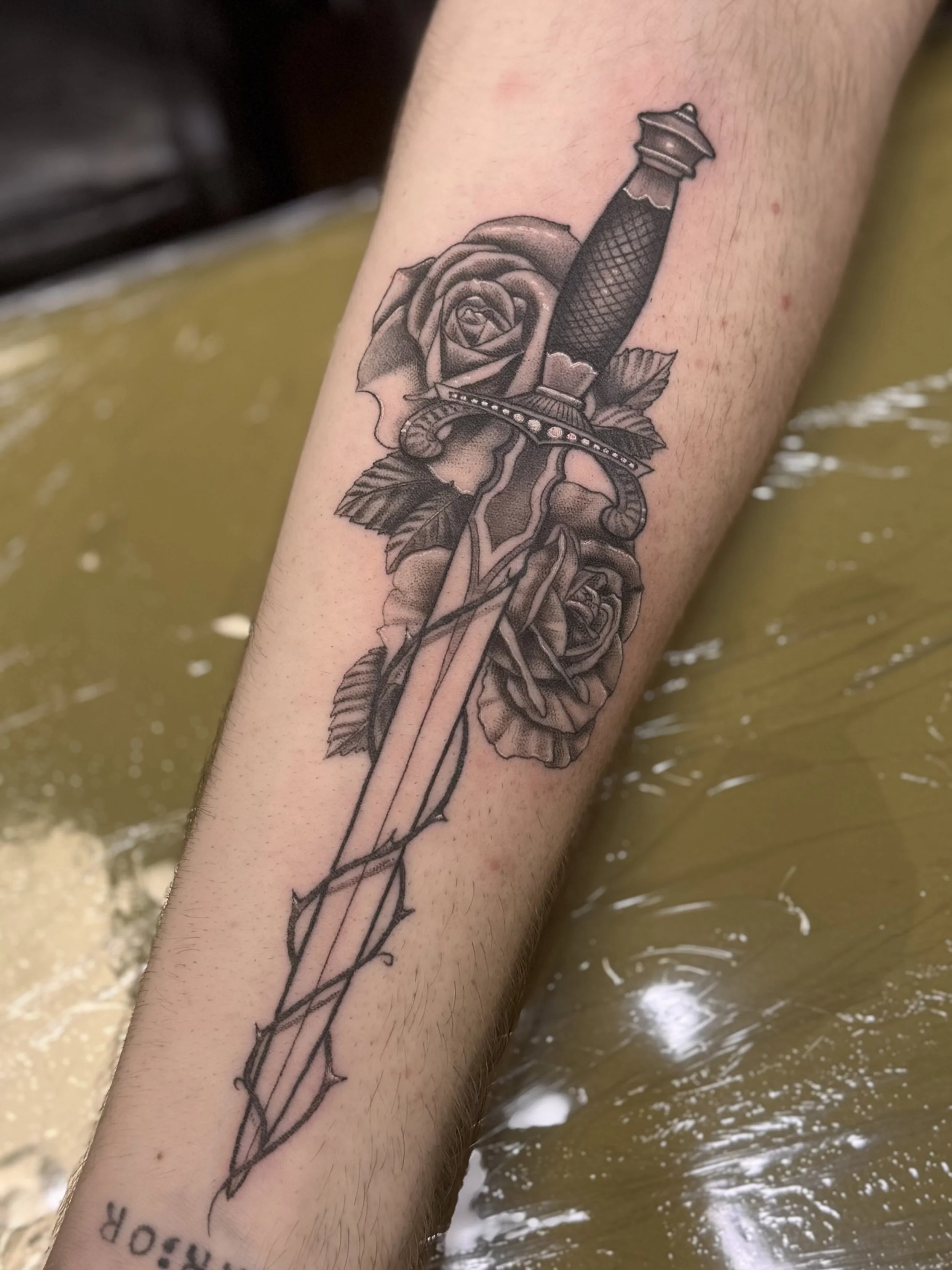 Tattoo of a rose and a dagger on a person's forearm, with water in the background.
