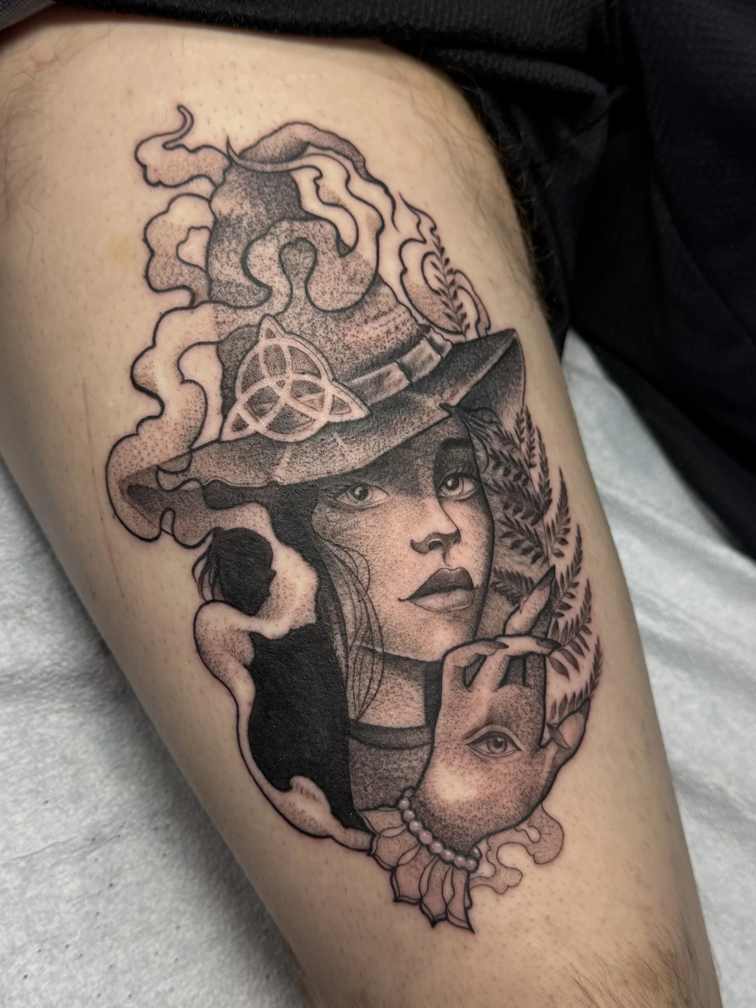 Tattoo of a woman’s face with her eyes looking sideways, a witch’s hat with a pentagram, smoky elements, leaves, a pearl necklace, and other decorative details on the arm.
