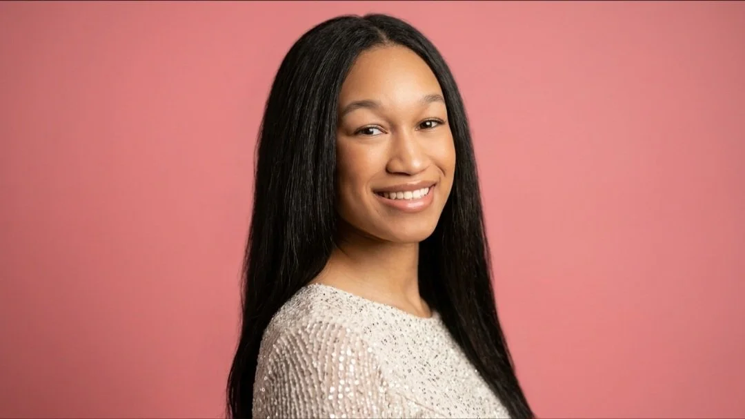 A young woman with long black hair, smiling, wearing a light-colored, shiny, textured top, standing against a pink background.