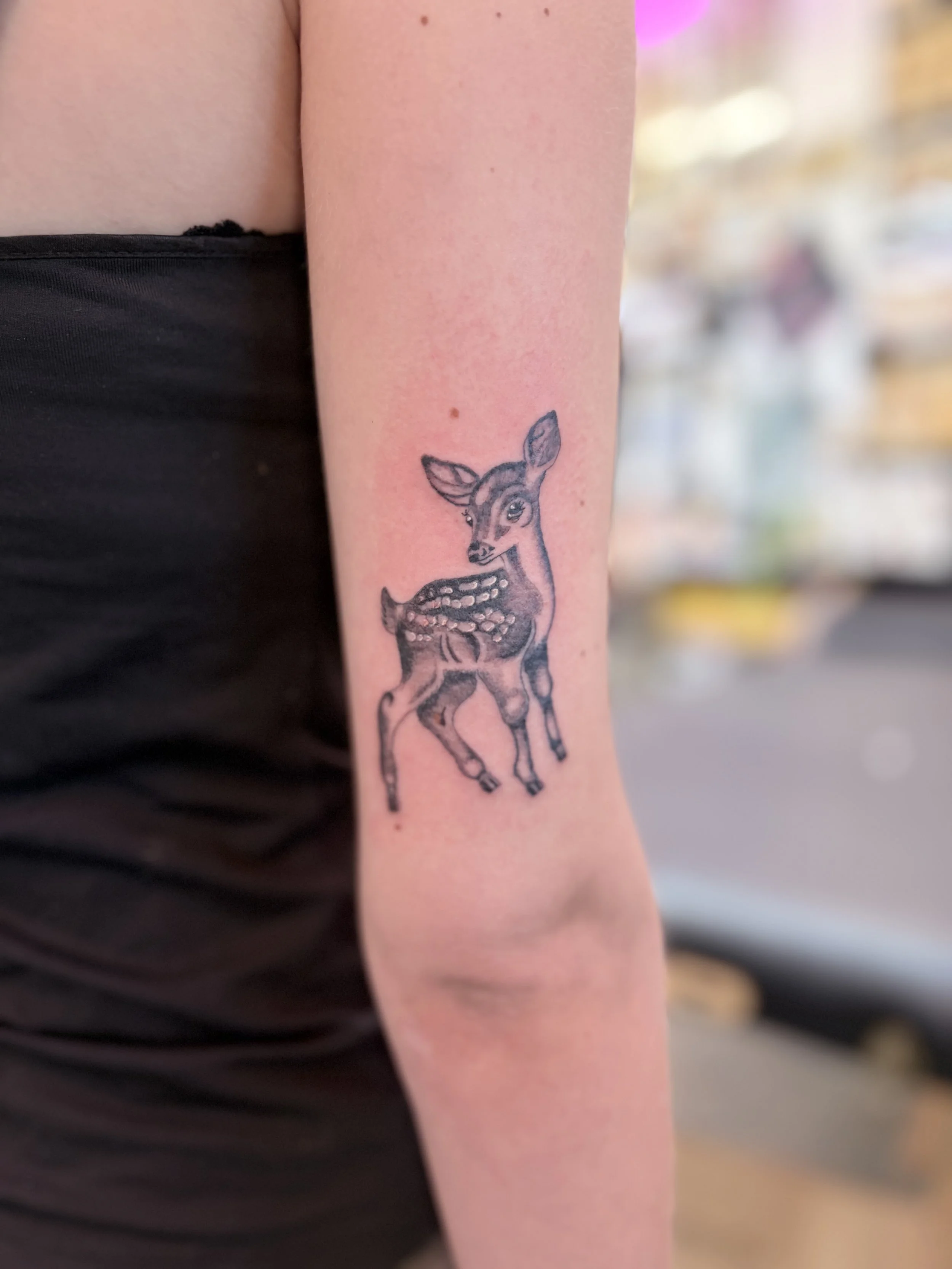 A person's upper arm with a black ink tattoo of a baby deer or fawn, showing its head, ears, and part of its body, on the inner arm, near the elbow.