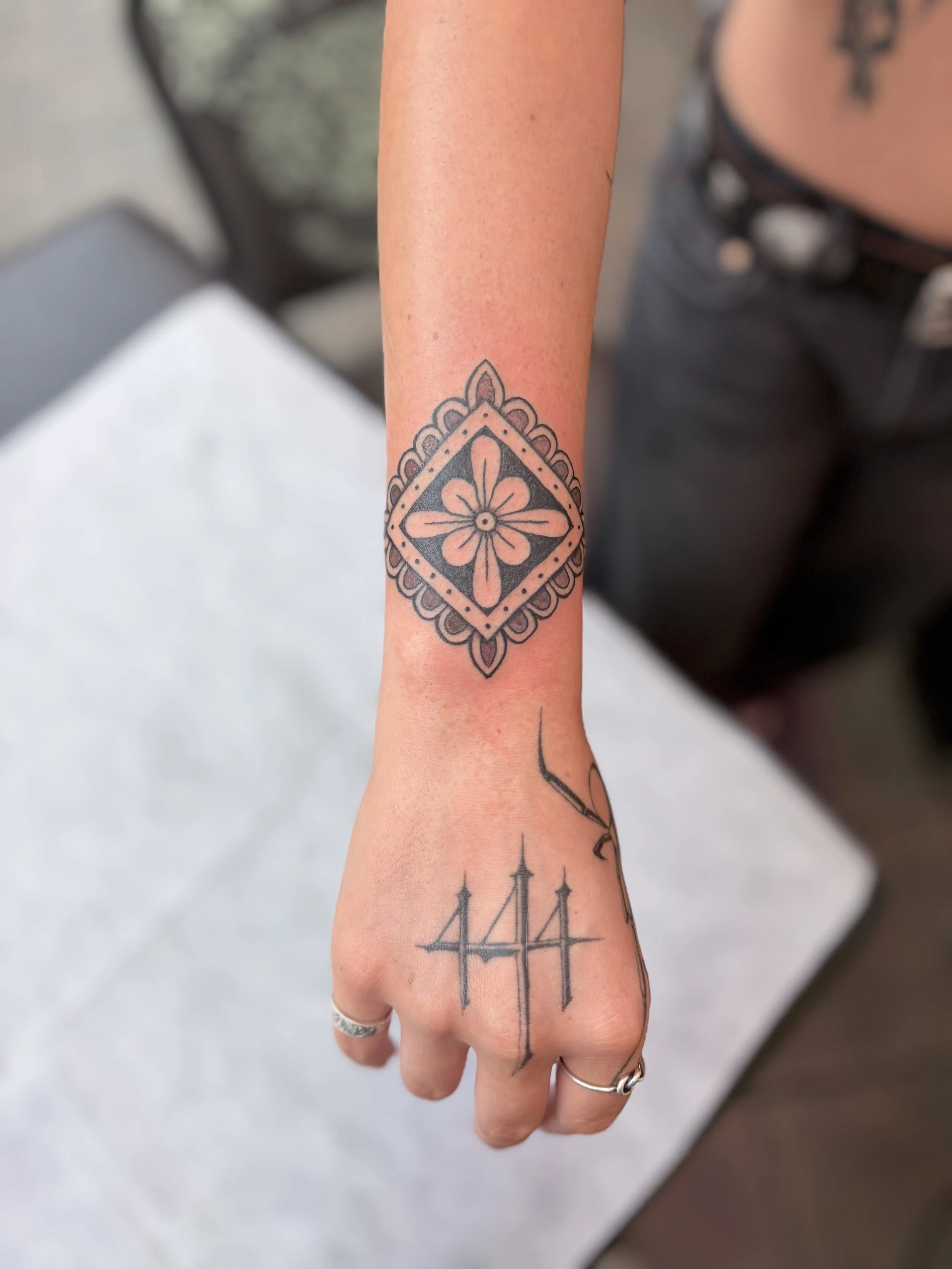 Close-up of a person's arm and hand with tattoos, including a black geometric floral design on the wrist and a line drawing of a ship on the forearm.