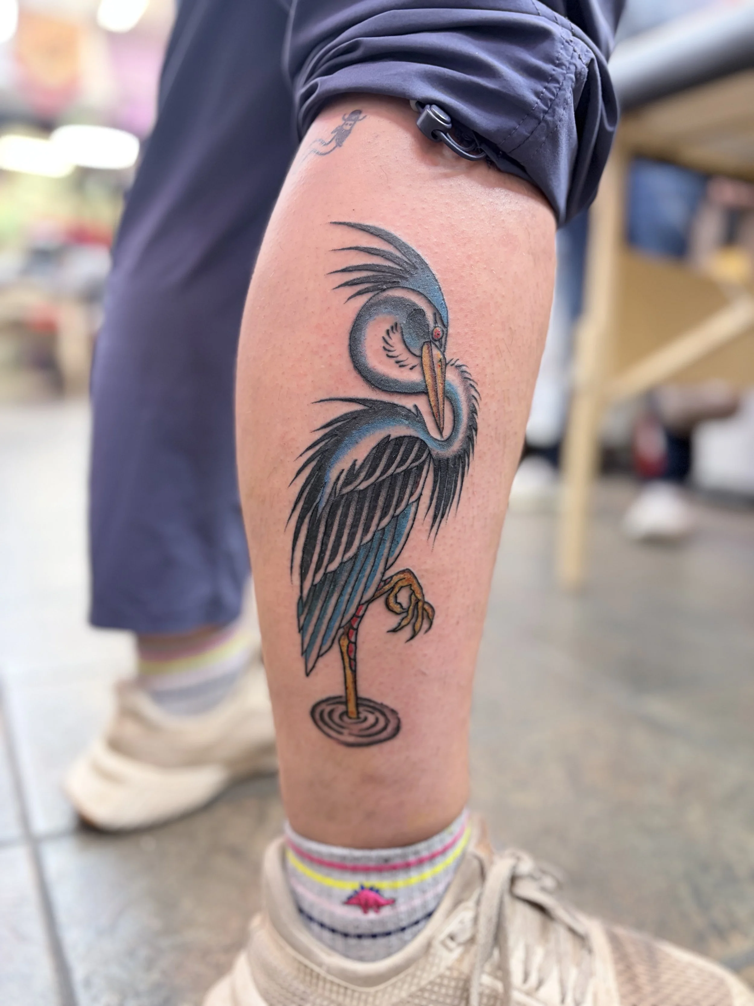A colorful tattoo of a heron or crane bird with blue, black, and orange ink on a person's upper leg, with the person wearing shorts and sneakers.