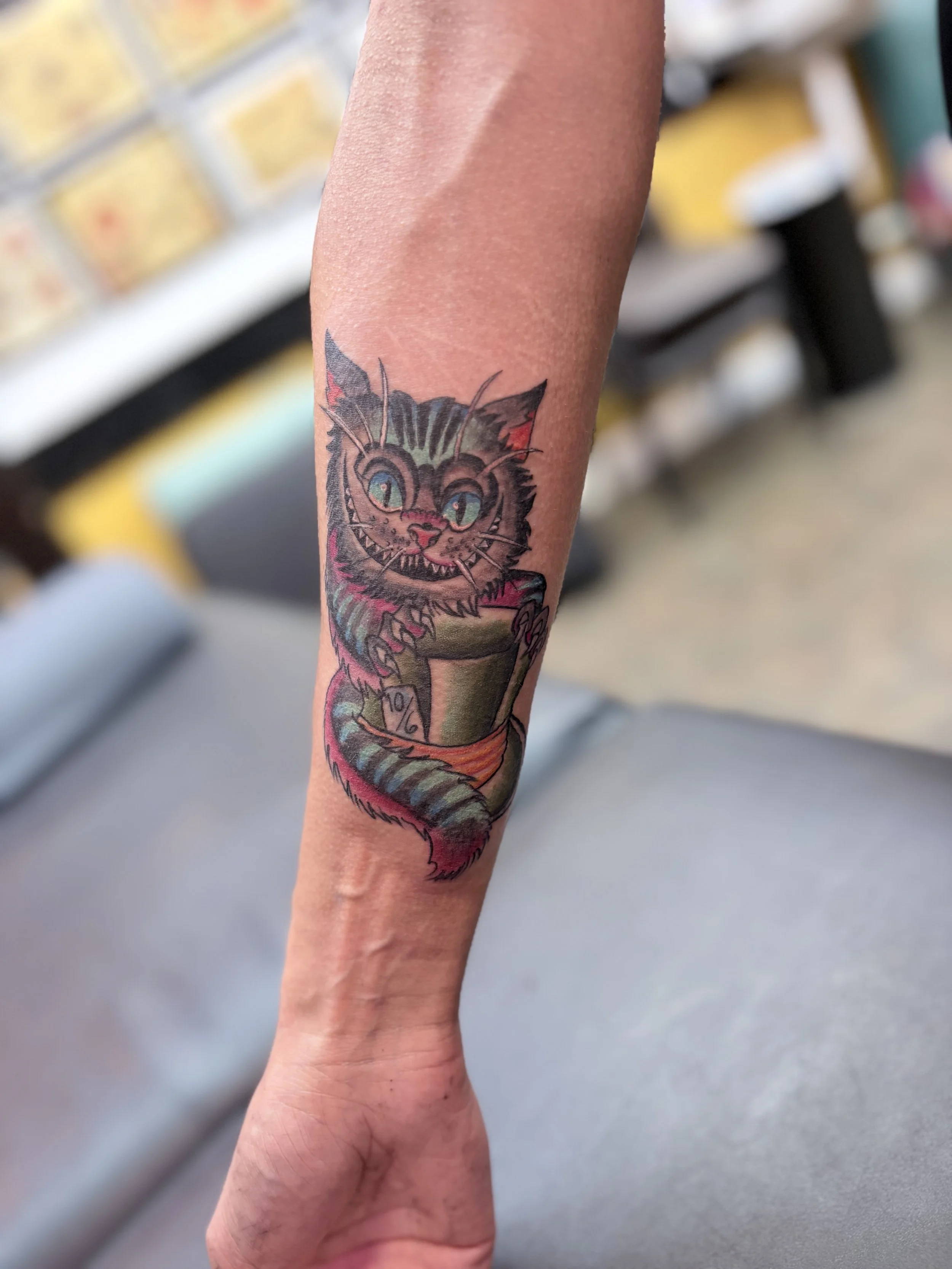 Colorful tattoo of a grinning cat with green eyes on a person's forearm, with a blurred background of a room with tiled walls and shelves.