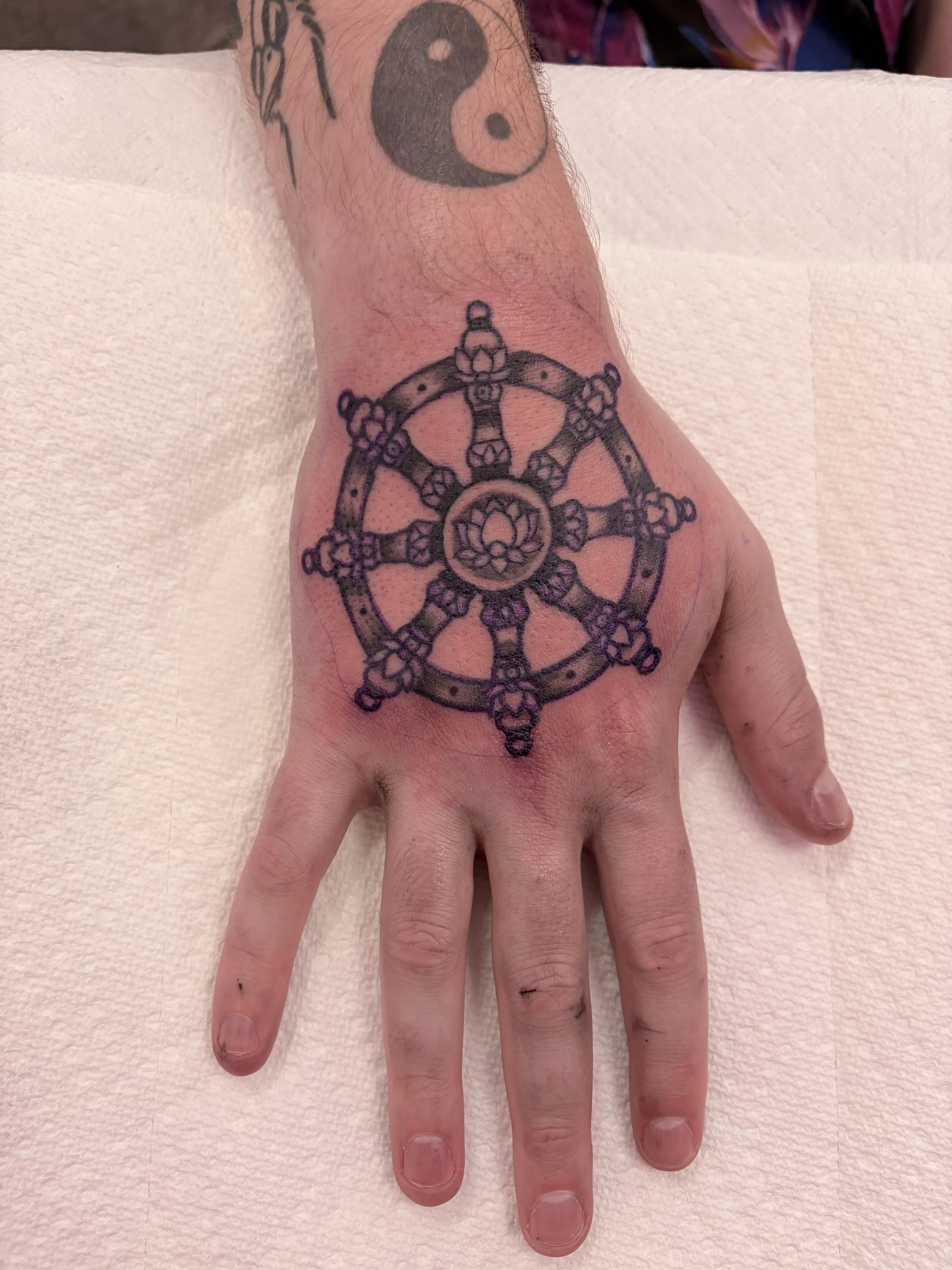 Tattoo of a Buddhist wheel with a lotus flower in the center on the person's hand. Additional tattoos partially visible on the wrist and forearm.