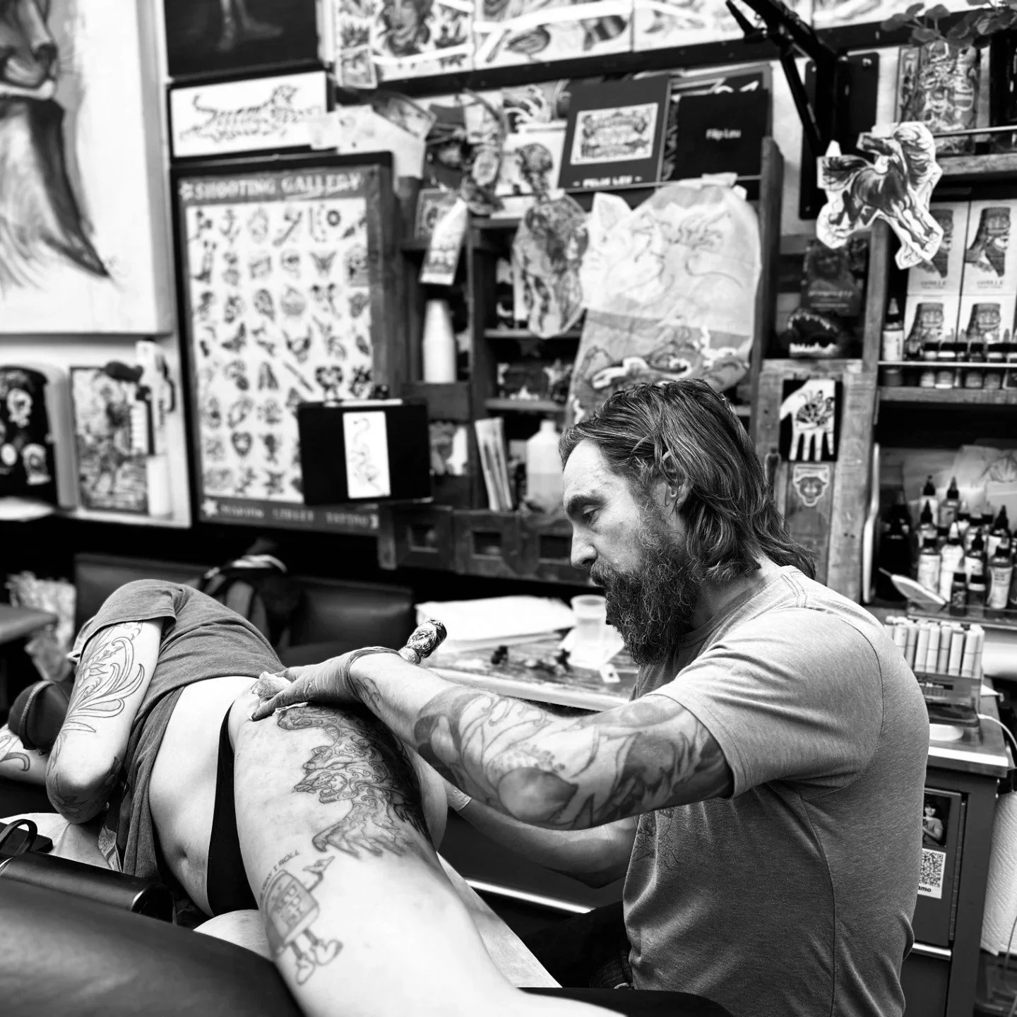 A man with long hair and a beard tattooing a person's arm in a tattoo shop, with shelves of art supplies and tattoo designs in the background.