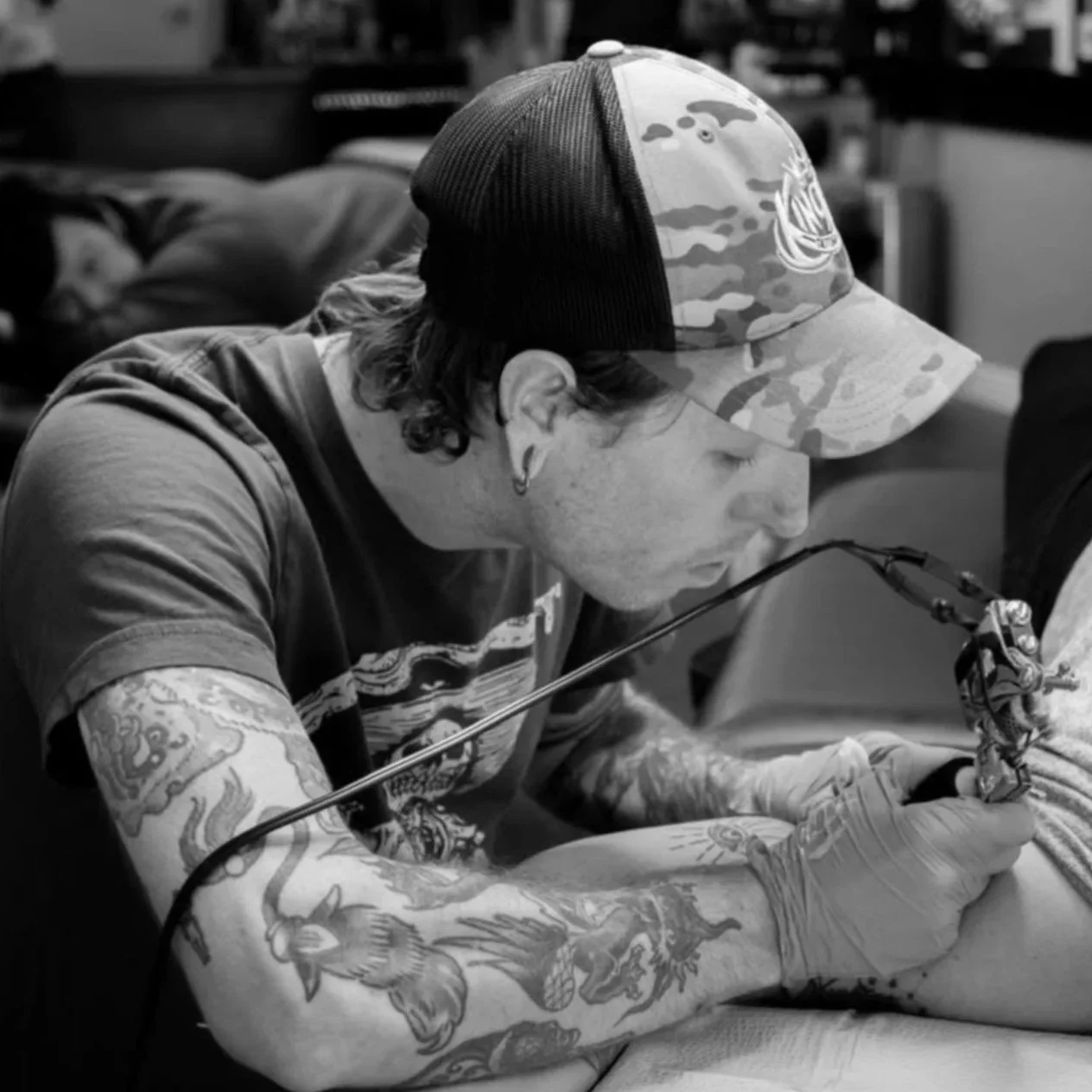 A person with tattoos on their arm and an earring, wearing a cap, tattooing or drawing on skin with a tattoo machine in a tattoo studio.