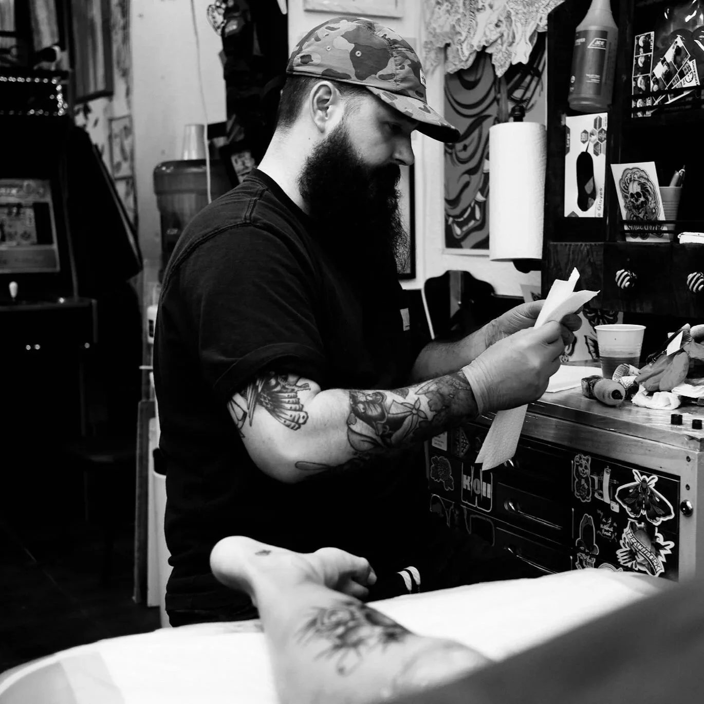 A man with a beard and tattoos on his arms, wearing a camo cap and black T-shirt, is standing and reading a piece of paper in a cluttered room decorated with artwork and stickers.