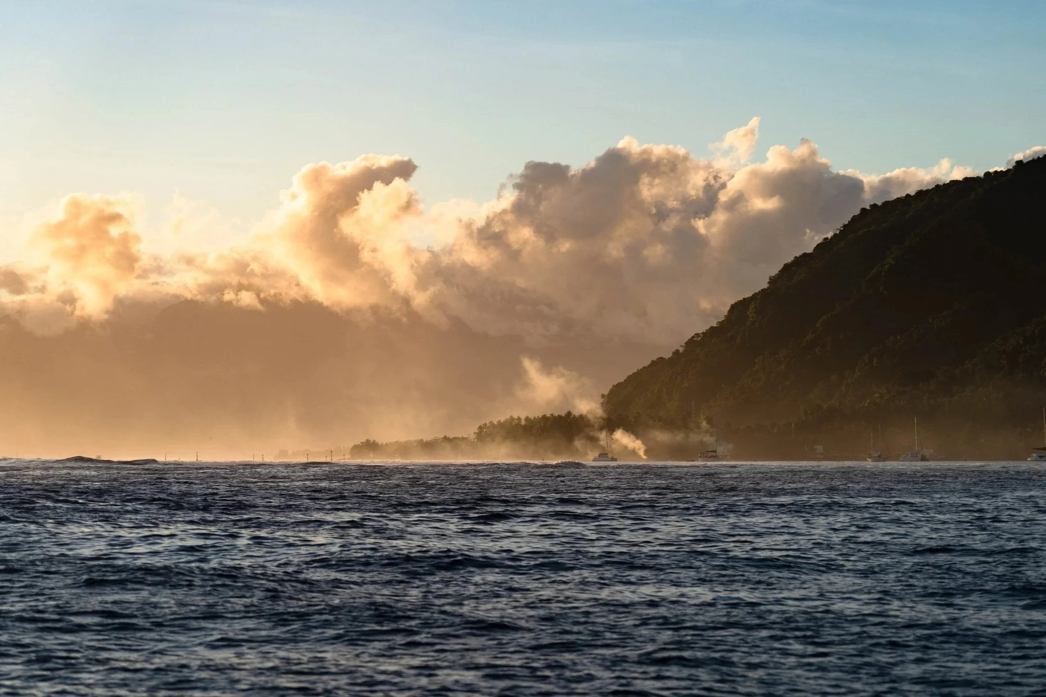 Ocean with waves, a hilly coastline covered in lush green trees, and a sky filled with large, fluffy clouds during sunset.