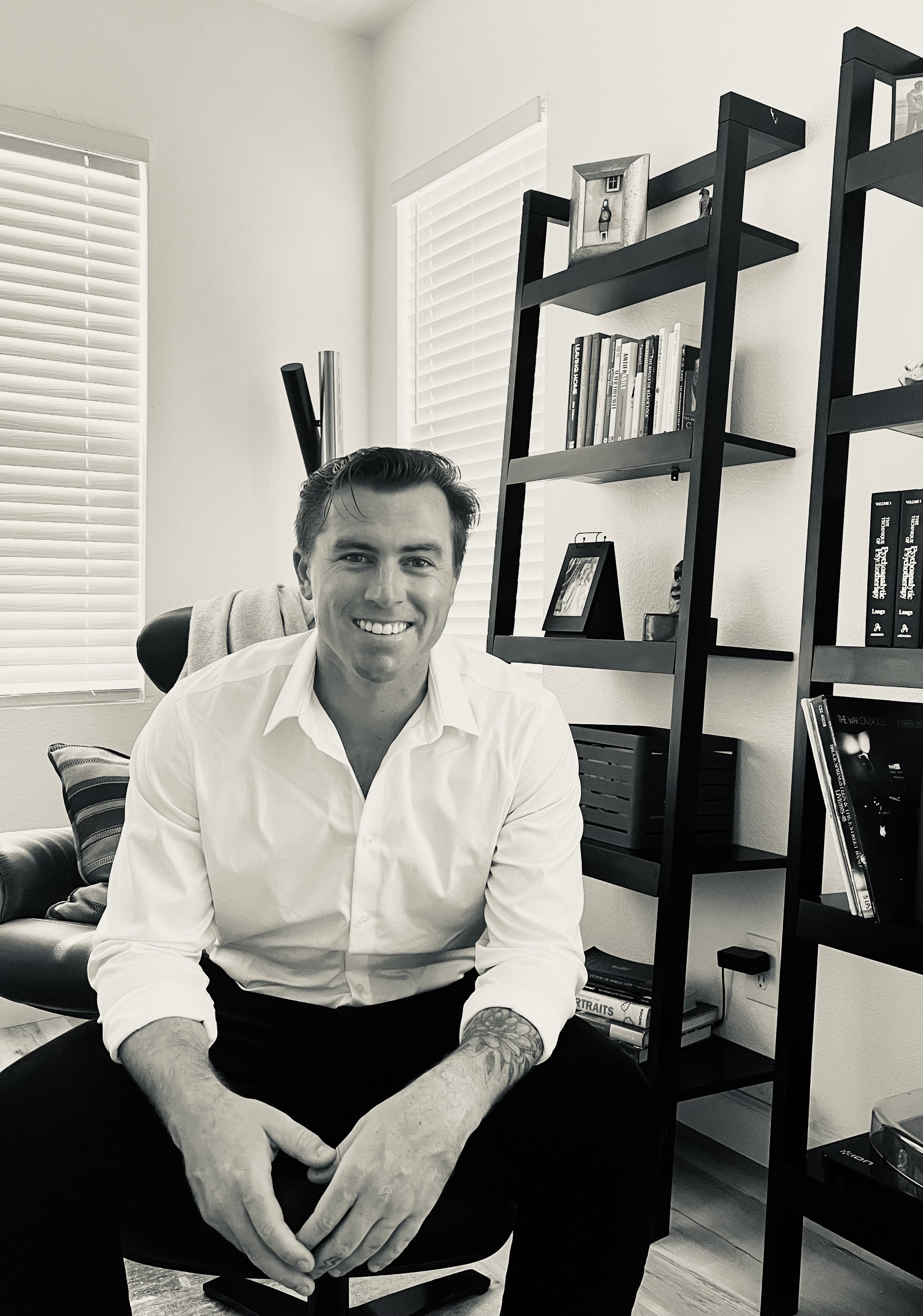 Black and white photo of a smiling man with dark hair, wearing a white shirt, sitting in an office or living room with a black bookshelf and window blinds in the background.