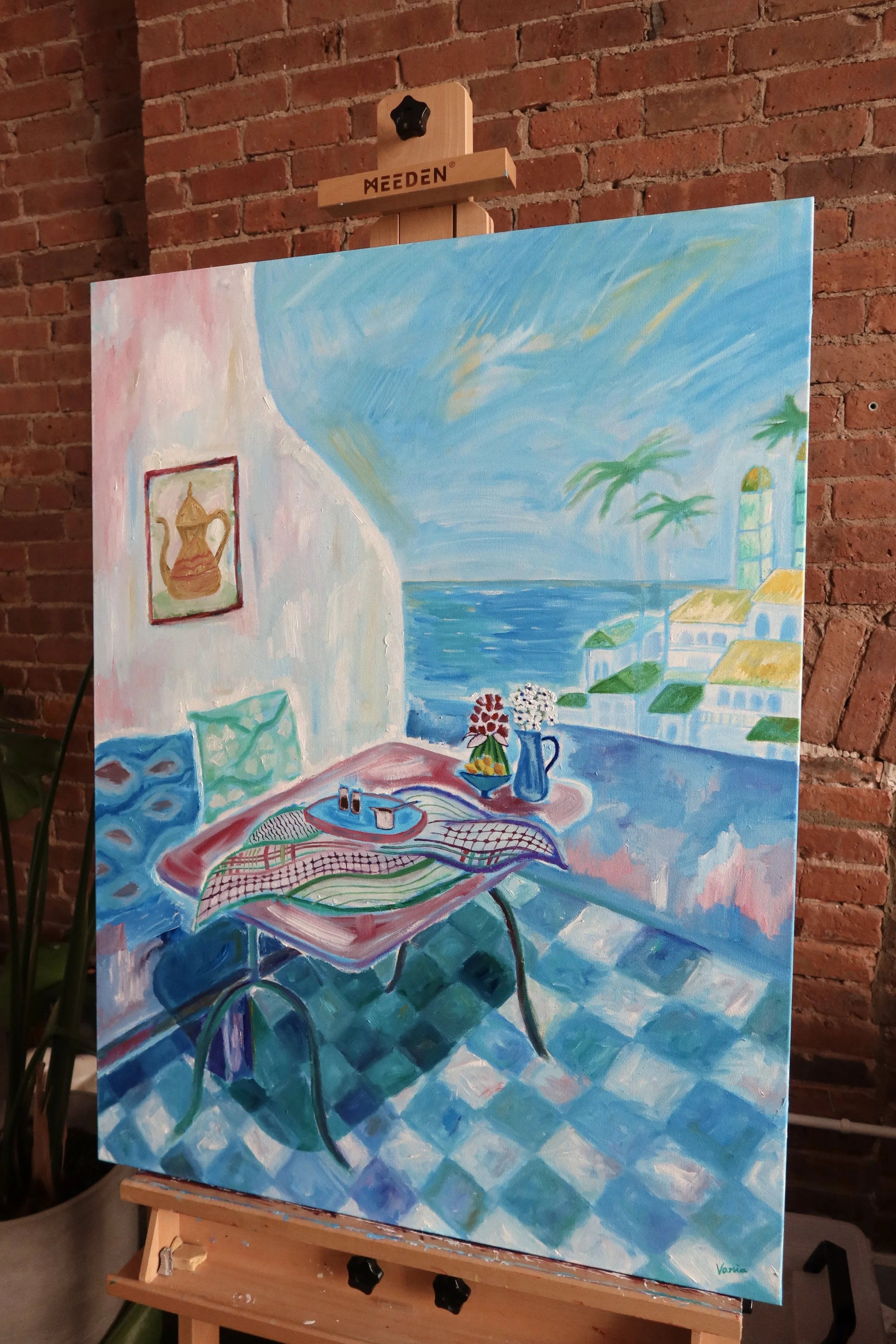 Painting of a colorful outdoor patio, with a table, chairs, flowers, and a view of the ocean and palm trees in the background, displayed on an easel against a brick wall.