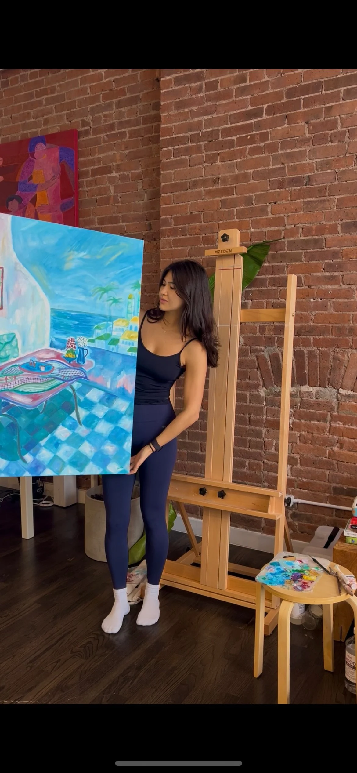 A young woman with dark hair wearing a black tank top and navy leggings standing indoors in front of an easel with a colorful painting of a seaside scene, with a brick wall in the background.
