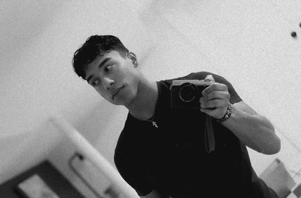 A young man taking a mirror selfie with a camera in a room, dressed in black, wearing a cross pendant, with short curly hair, in black and white.