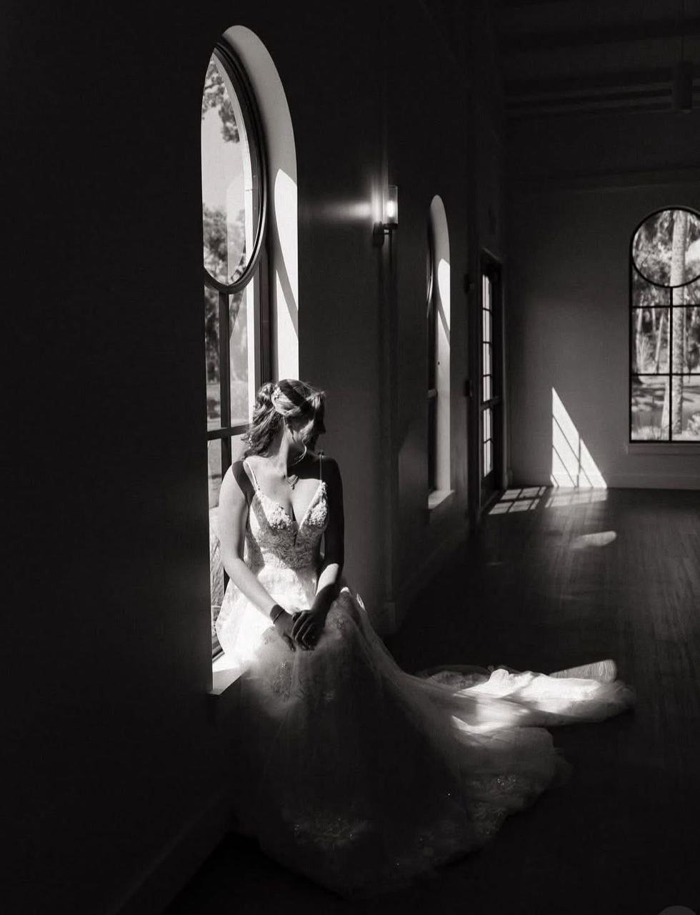 A woman in a wedding dress sitting by a large arched window with sunlight streaming in, creating shadows inside a room.