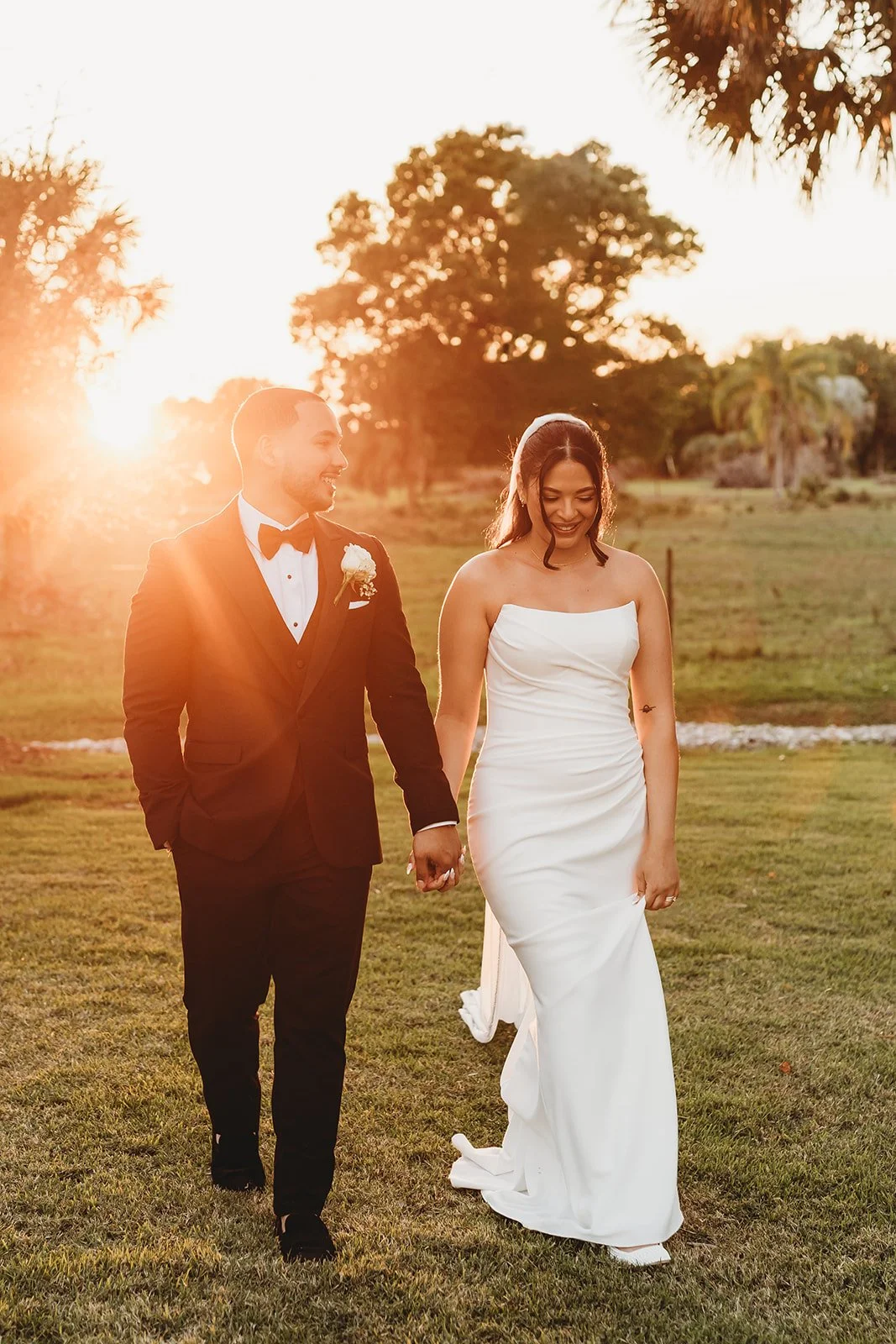 A newlywed couple walking hand in hand outdoors during sunset, with the groom in a black tuxedo and the bride in a strapless white wedding gown, surrounded by trees and grass.
