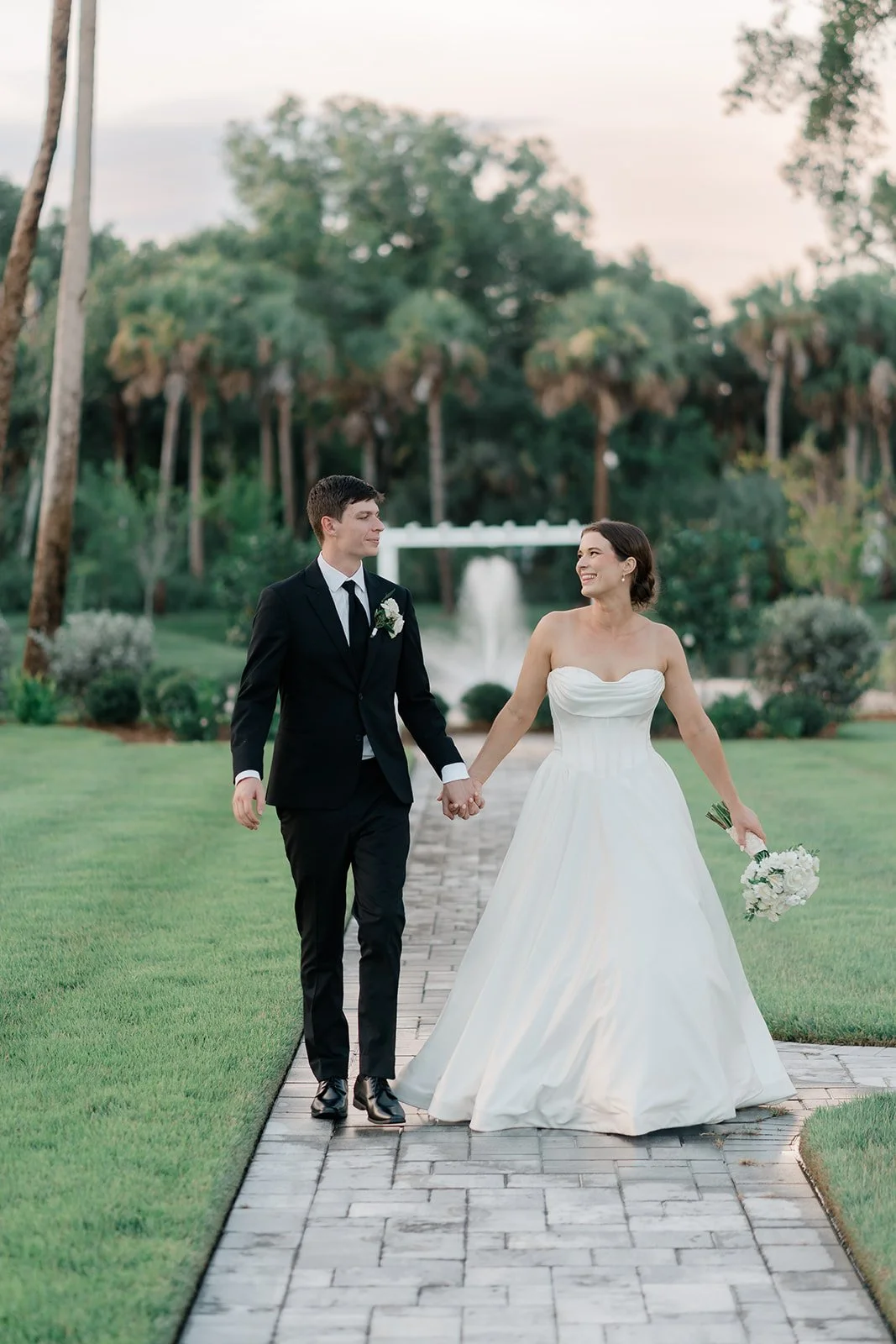 A bride and groom holding hands and walking outdoors on a stone path in a lush green garden with palm trees and a fountain in the background during sunset.